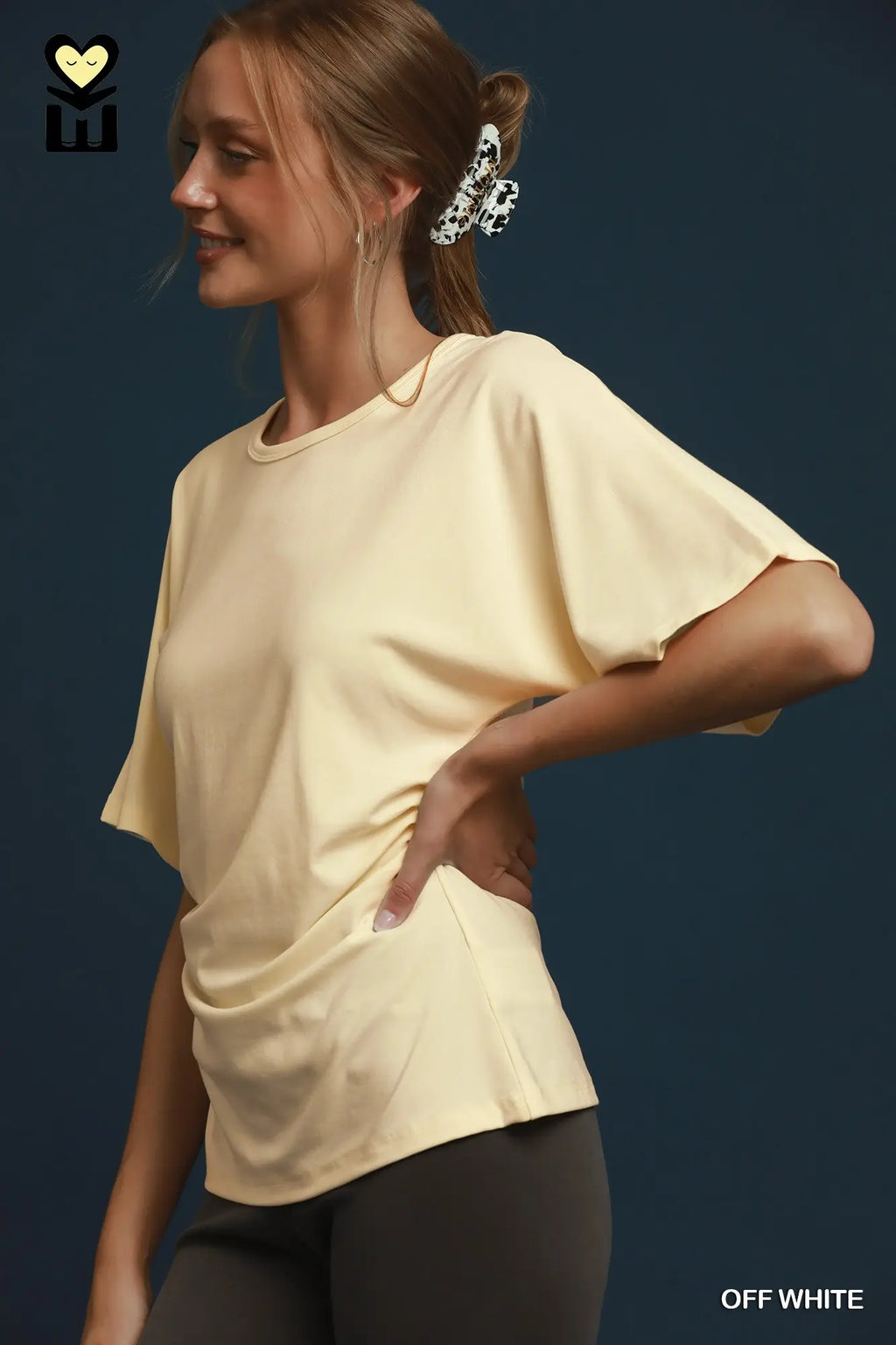 A woman wearing a short-sleeve off-white t-shirt with a draped front detail, paired with dark gray leggings, posed against a dark blue background. The brand logo 'EW' is visible in the top-left corner, and 'OFF WHITE' is written in the bottom-right corner.