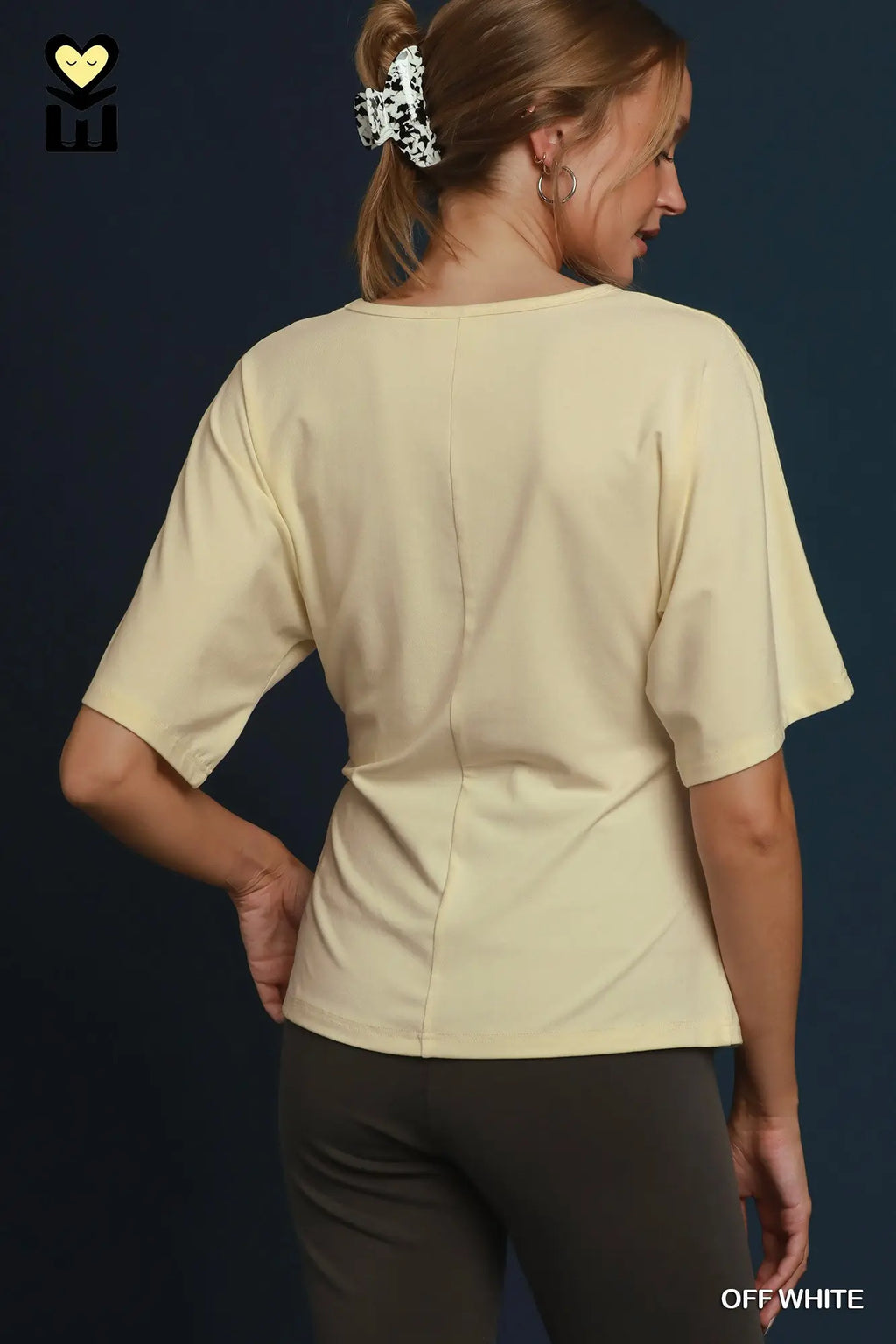 A woman shown from the back wearing a cream-colored short-sleeve top and dark gray pants against a dark navy background. The top has a subtle vertical seam down the center back.