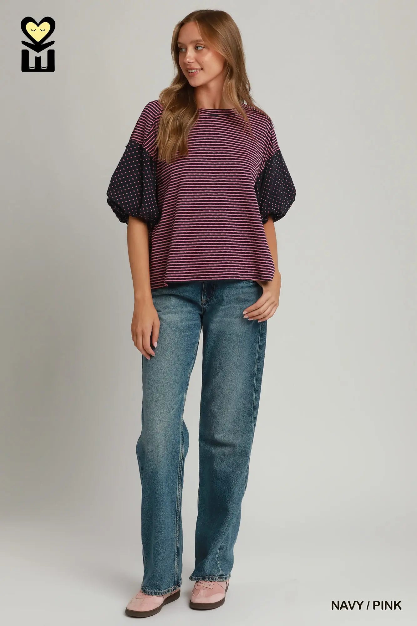 A woman modeling a short-sleeve knit top with horizontal navy and pink stripes on the body and solid navy polka-dot sleeves, paired with blue jeans and pink shoes, against a light gray studio background.