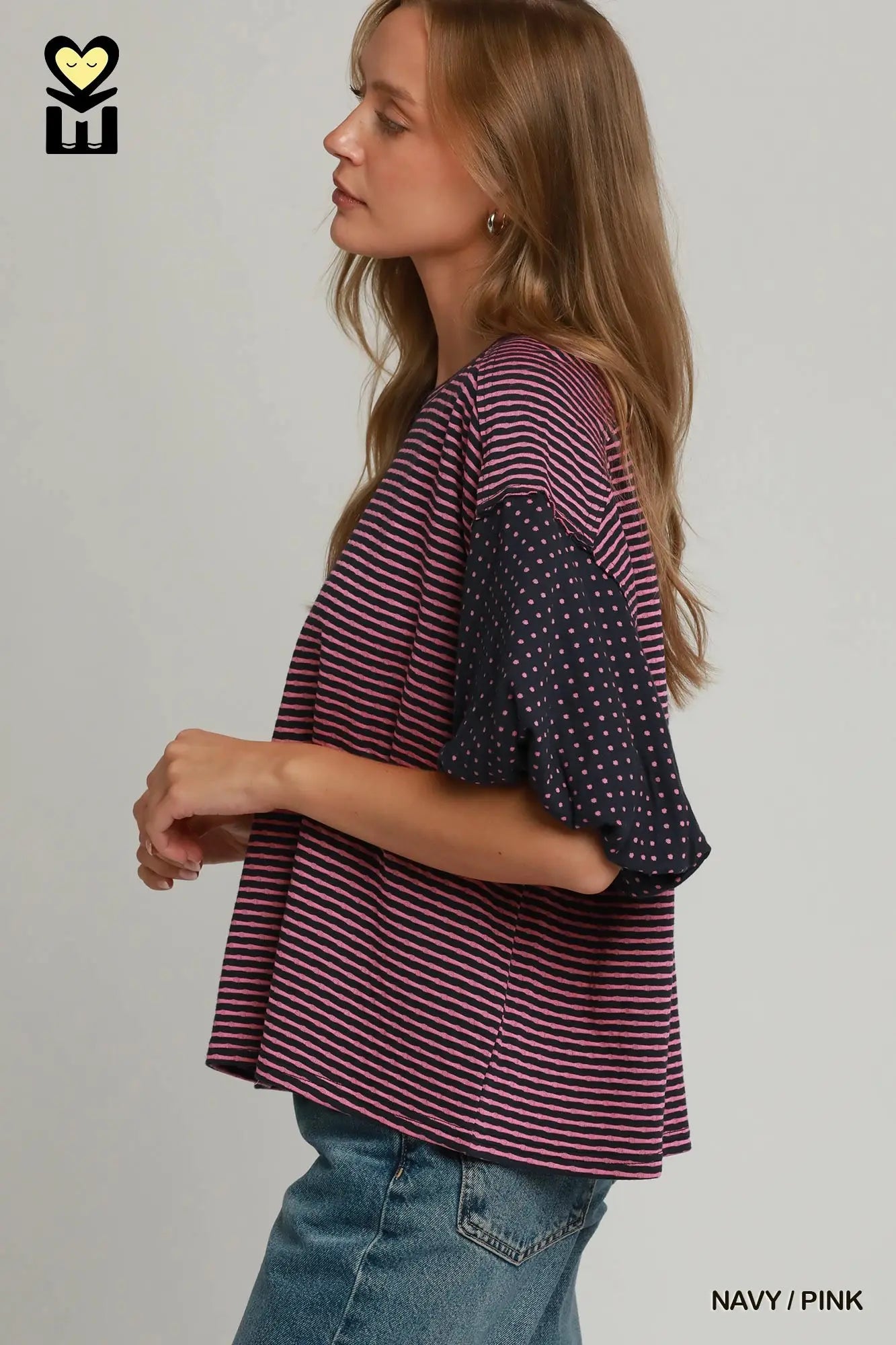 A woman shown in side profile wearing a loose short-sleeve top with pink and navy horizontal stripes on the main body and solid navy flutter sleeves with small white polka dots. She is also wearing blue jeans against a plain light gray studio background.