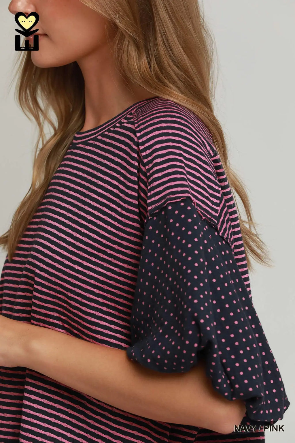 A close-up of a woman wearing a striped top with dark navy sleeves featuring small pink polka dots. The top has thin horizontal stripes in pink and navy across the body.