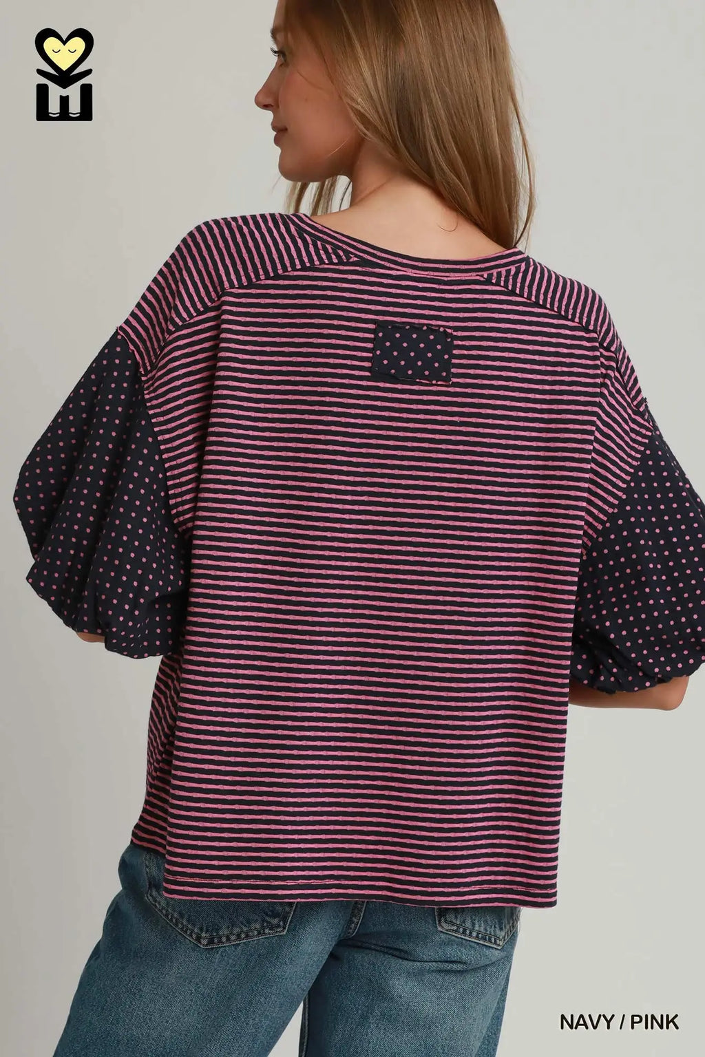 A woman shown from the back wearing a loose striped top with navy polka-dot puff sleeves and a small navy patch on the upper back, paired with blue jeans against a light gray studio background.