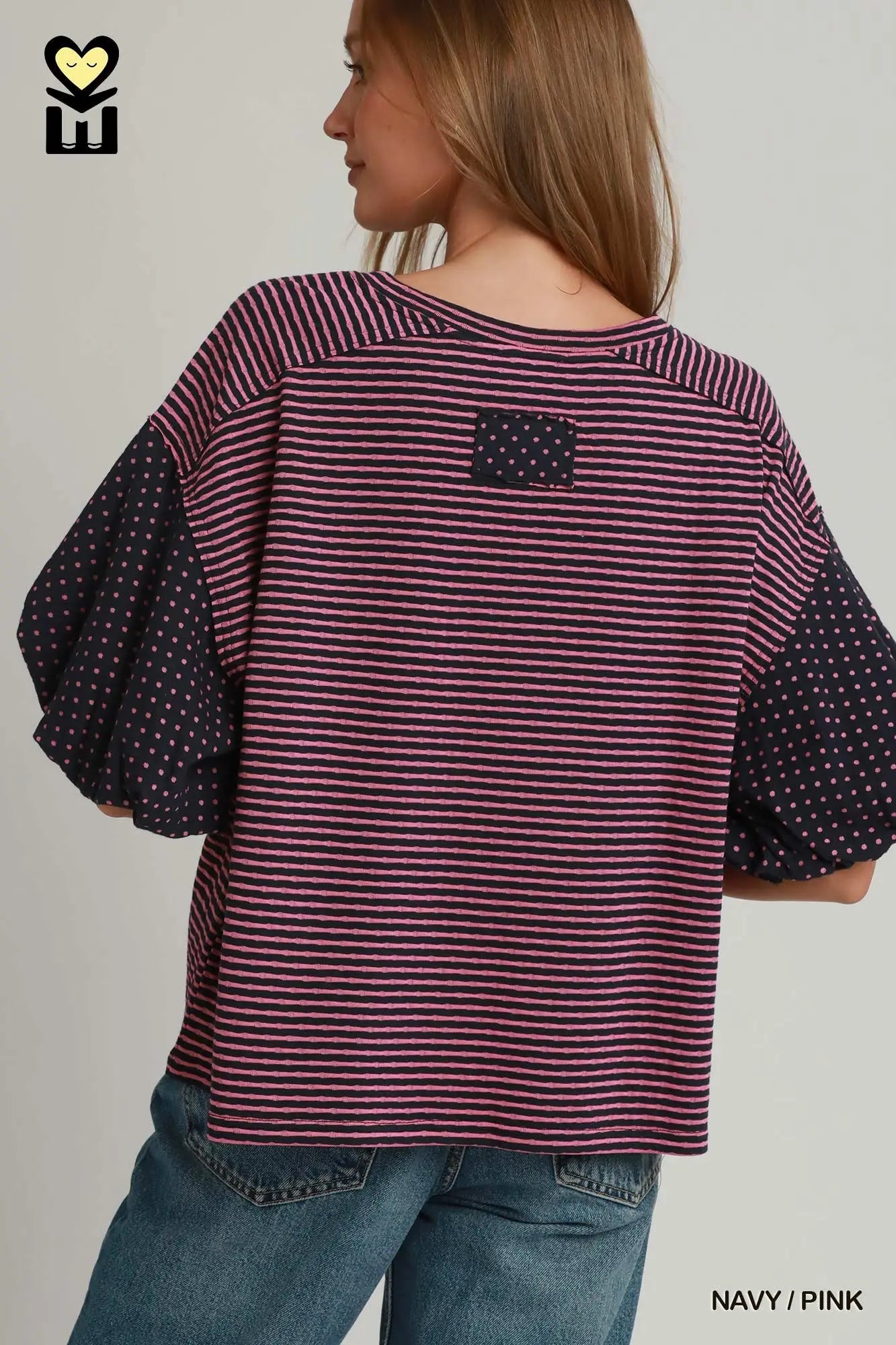 A woman shown from the back wearing a loose striped top with navy polka-dot puff sleeves and a small navy patch on the upper back, paired with blue jeans against a light gray studio background.