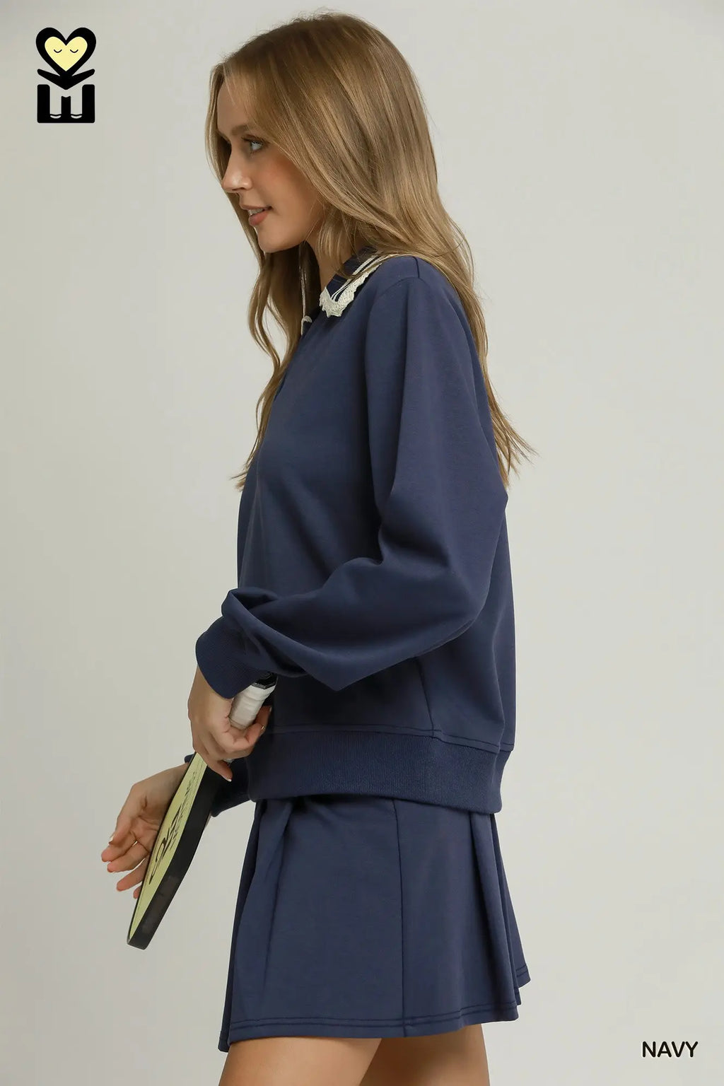 A woman modeling a navy blue sweatshirt and matching pleated mini skirt set against a plain light gray studio background. She is shown in profile holding a small clutch, with a small 'NAVY' text label in the bottom right corner and a logo mark in the top left.