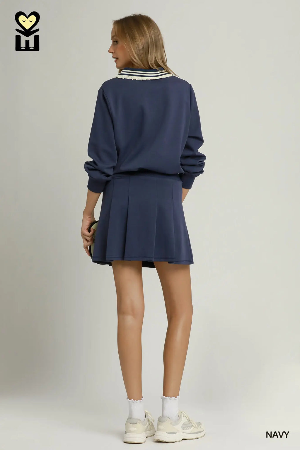 A model is shown from the back wearing a navy blue sweatshirt and matching pleated mini skirt set. The sweatshirt features a white and navy striped collar.