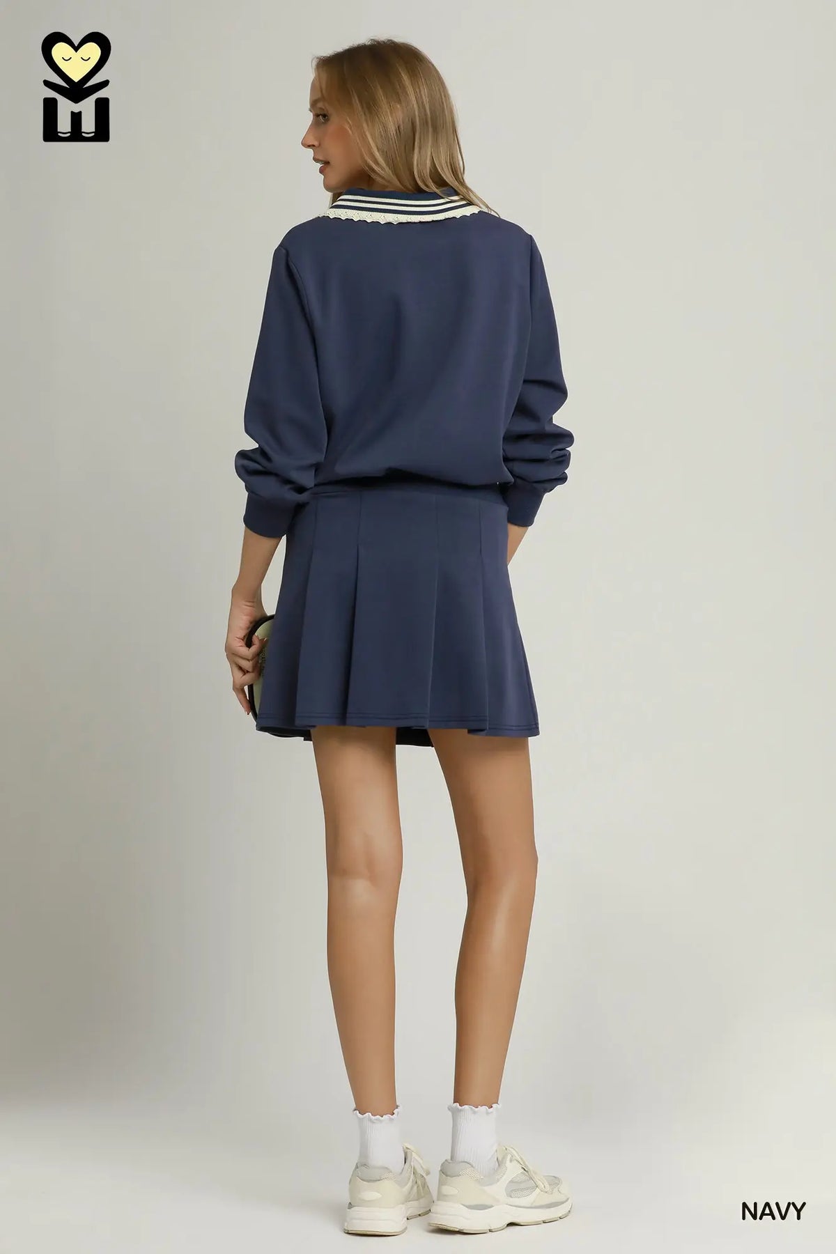 A model is shown from the back wearing a navy blue sweatshirt and matching pleated mini skirt set. The sweatshirt features a white and navy striped collar.