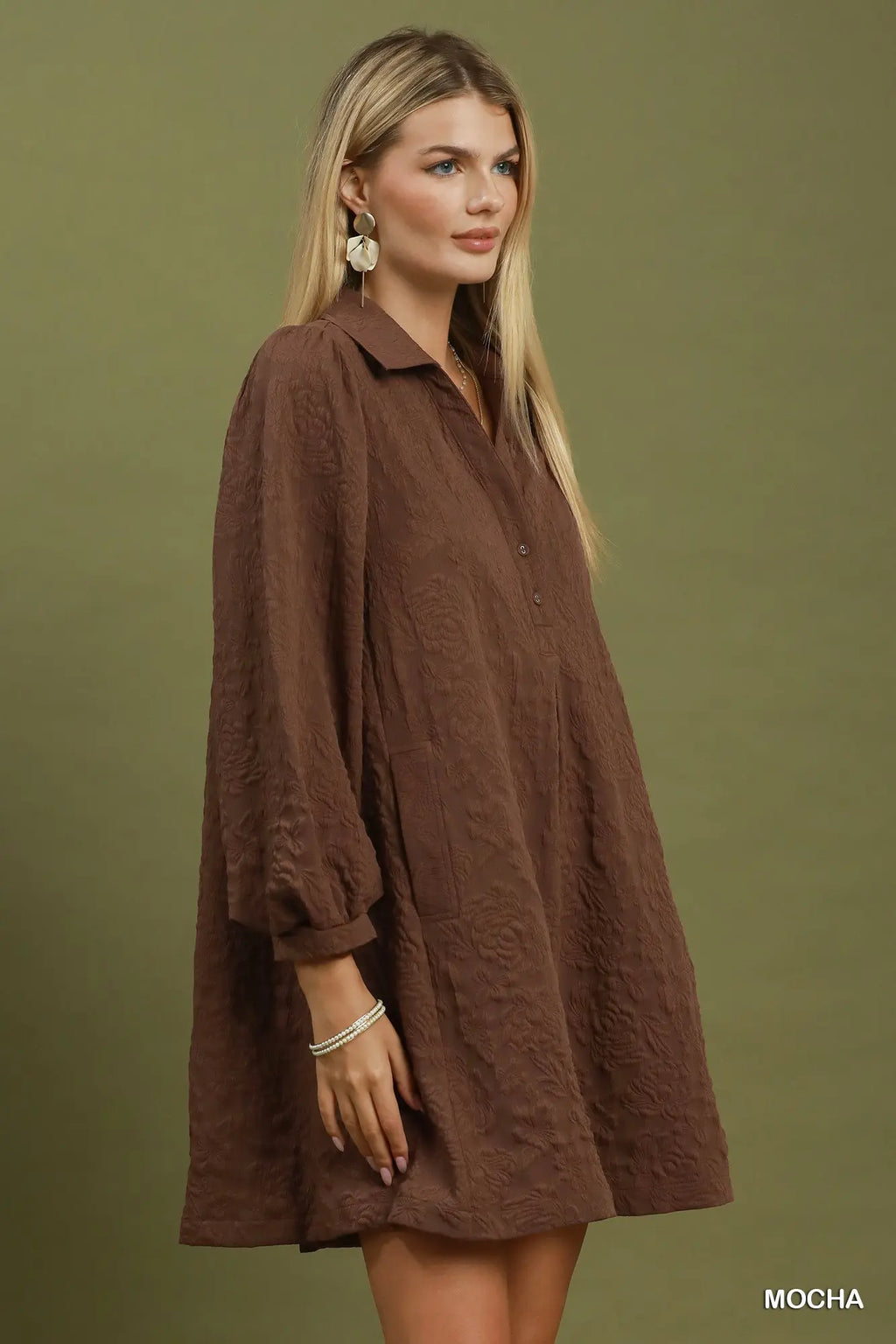 A woman modeling a brown textured long-sleeve shirt dress against a solid olive green studio background. The dress has a relaxed, slightly oversized fit with a collar and button-down front.