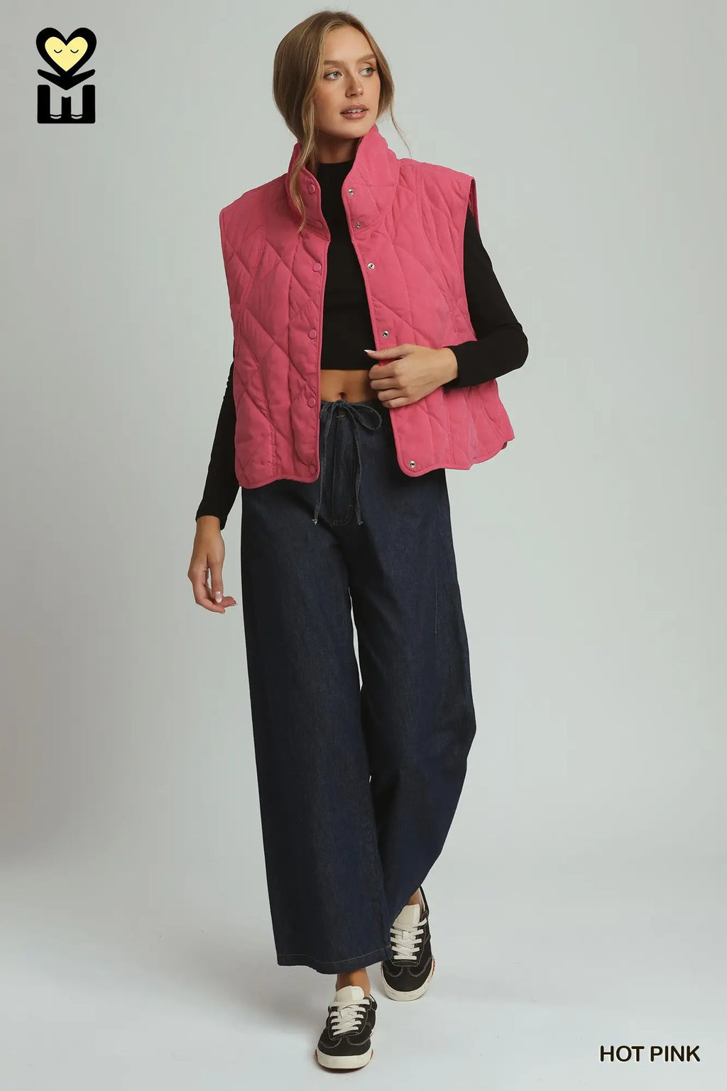 A model wearing a hot pink quilted sleeveless puffer vest over a black long-sleeve top, paired with dark wide-leg pants and black sneakers, against a light gray studio background.