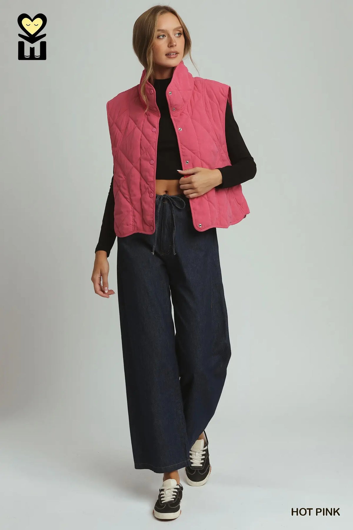 A model wearing a hot pink quilted sleeveless puffer vest over a black long-sleeve top, paired with dark wide-leg pants and black sneakers, against a light gray studio background.