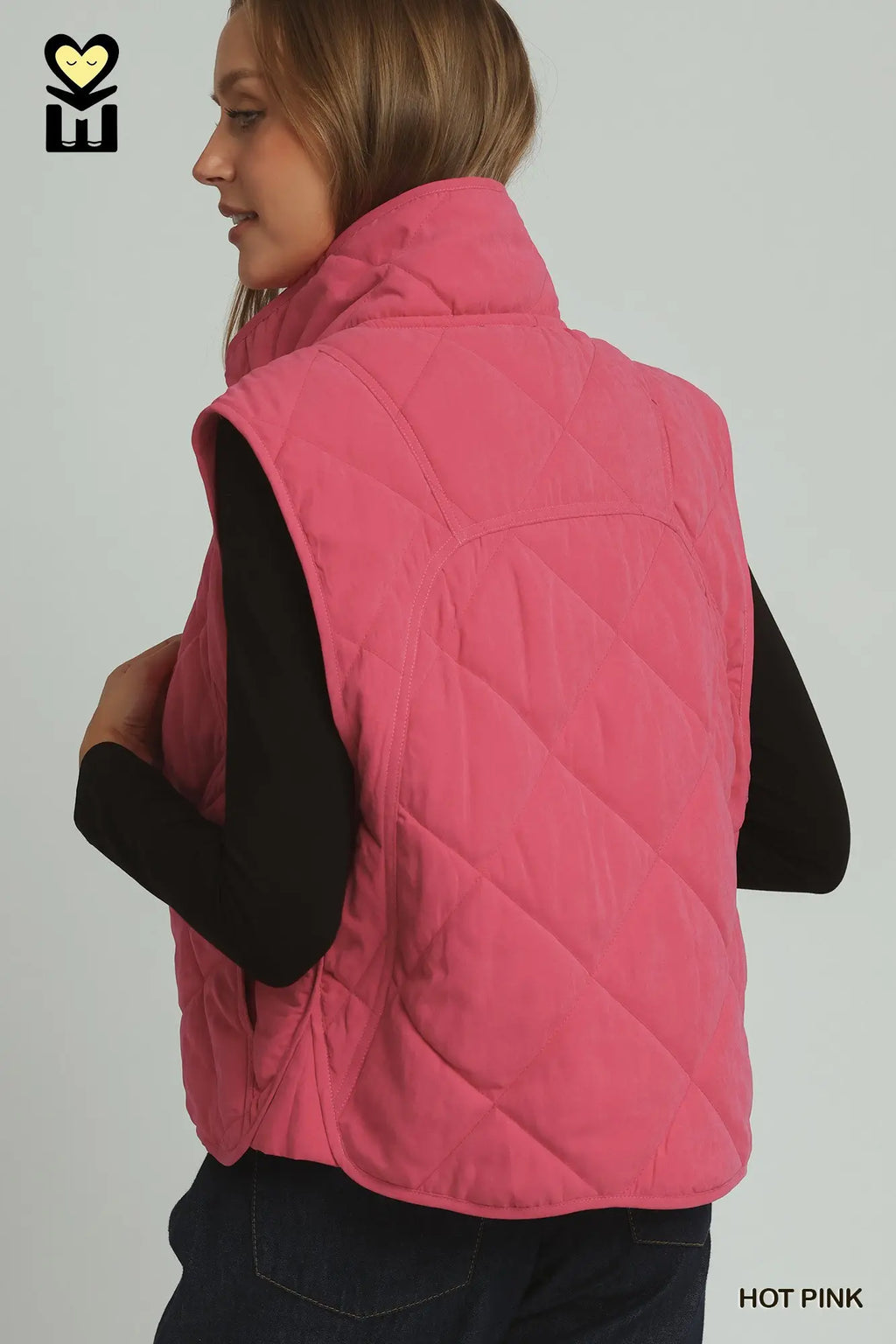 A woman shown from the back wearing a quilted sleeveless puffer vest in bright pink over a black long-sleeve top and dark jeans, against a light gray studio background. The text 'HOT PINK' appears in the lower right corner.