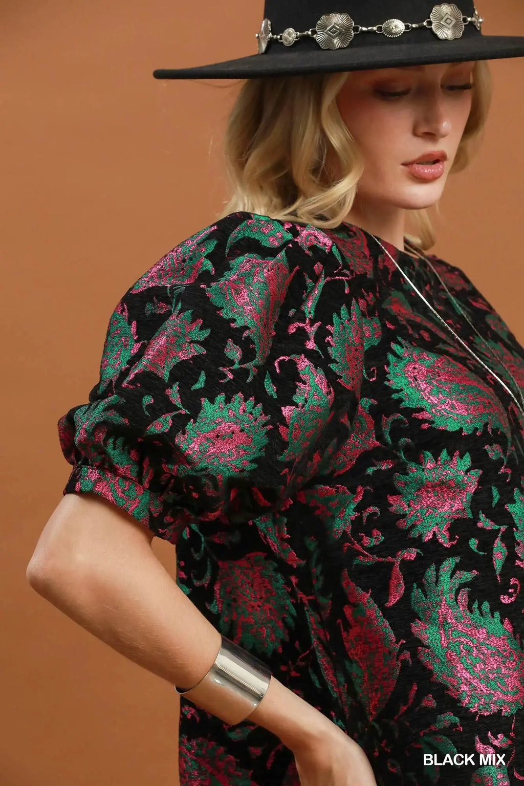A woman wearing a black patterned blouse with green and pink paisley motifs, a black wide-brim hat with silver conchos, and a silver cuff bracelet, posed against a solid warm brown background.