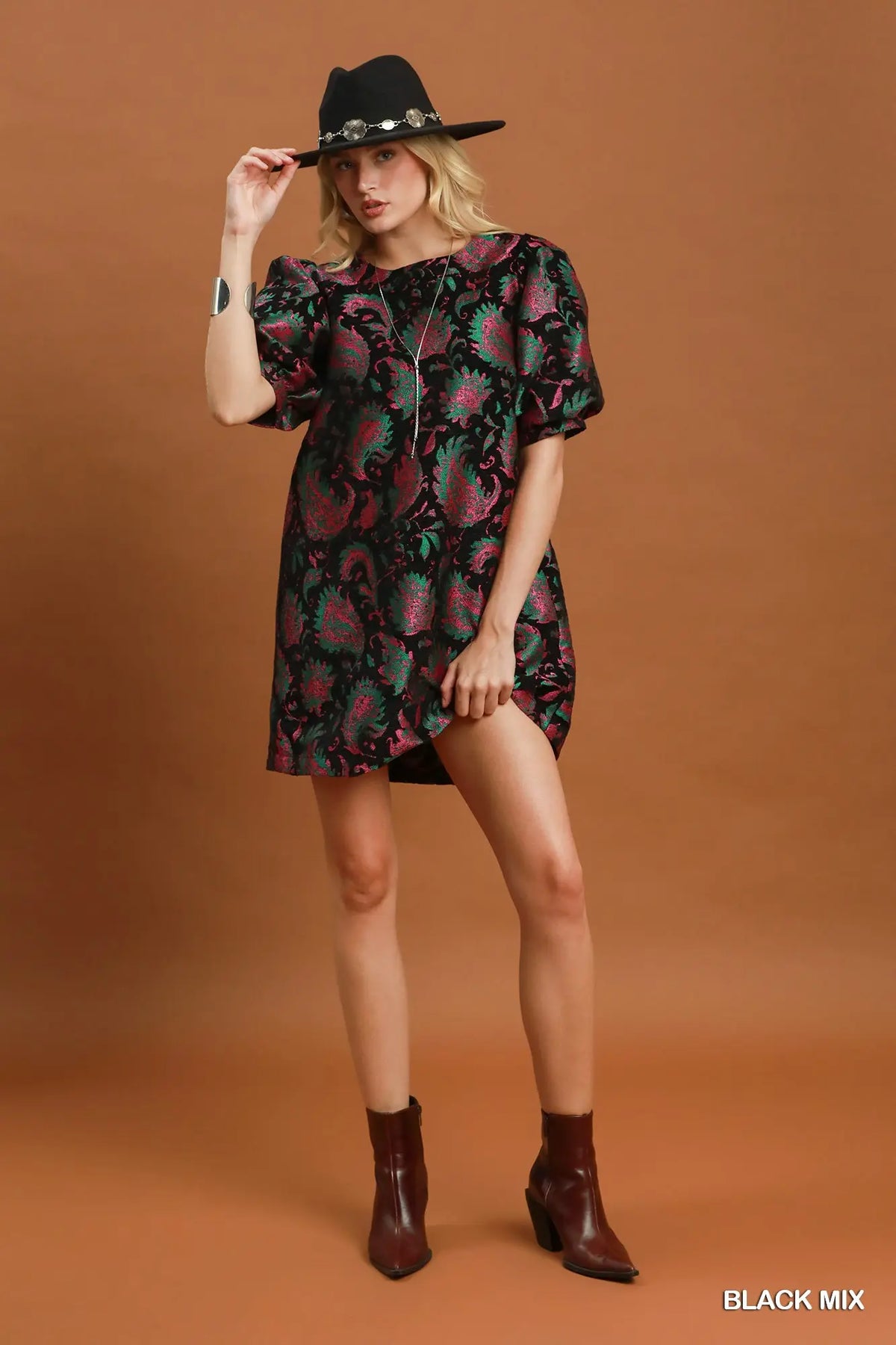 A woman modeling a short black dress with a vibrant pink and green paisley pattern, styled with a black hat and brown ankle boots against a solid brown studio background.