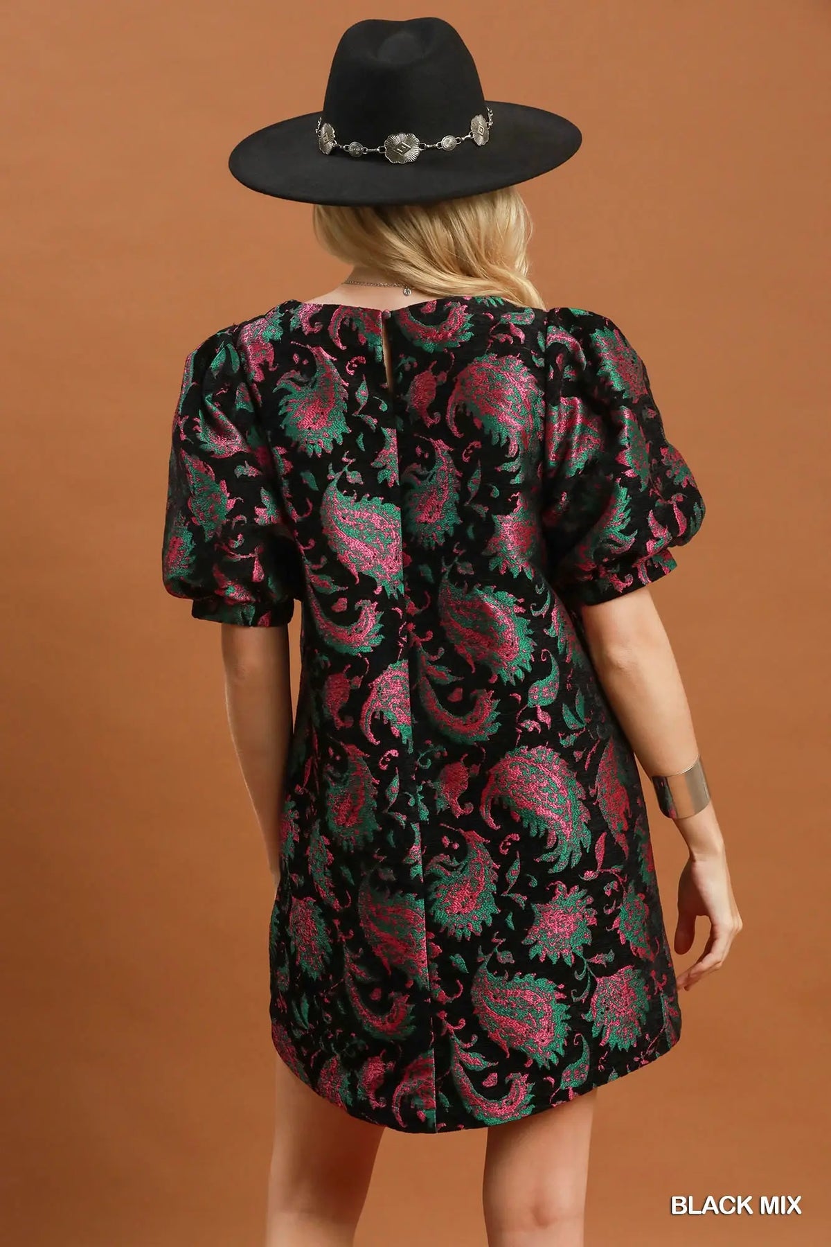 A woman shown from the back wearing a short puff-sleeve dress with a black, pink, and green paisley pattern, paired with a black wide-brim hat, against a solid brown background.