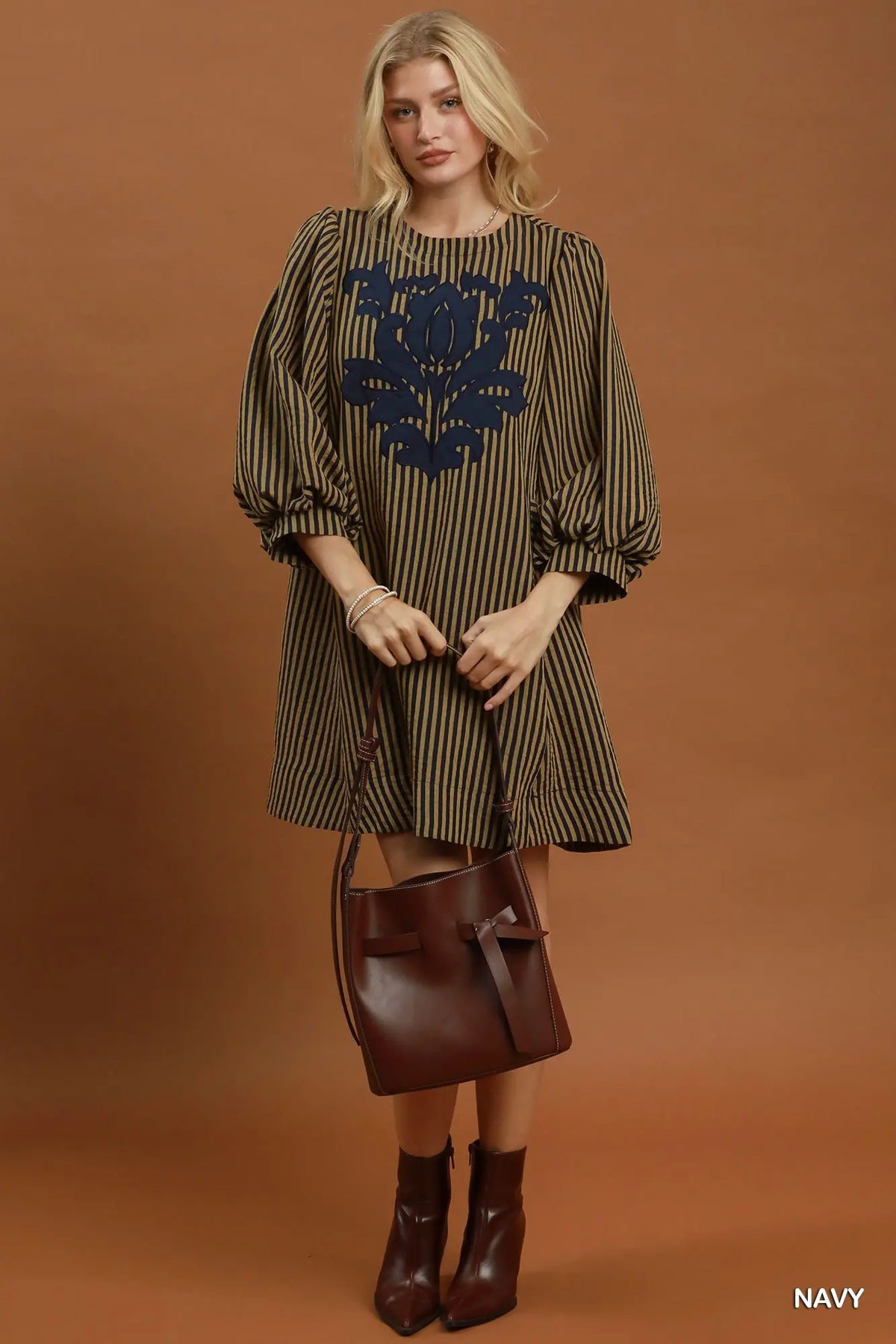 A woman models a brown and black vertically striped tunic dress with a navy blue embroidered floral pattern on the chest. She is holding a brown leather bucket bag and wearing brown leather ankle boots, set against a solid brown studio background.