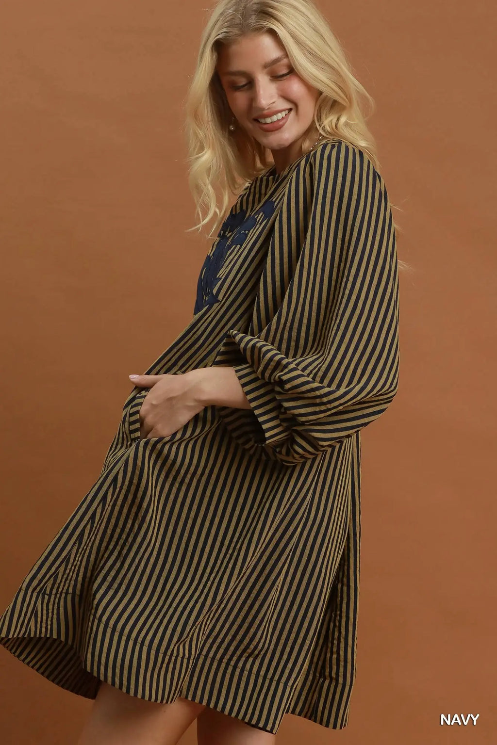 A woman wearing a loose, knee-length striped dress with wide sleeves and a navy blue underlayer, posed against a solid warm brown studio backdrop. The word 'NAVY' appears in the bottom right corner.