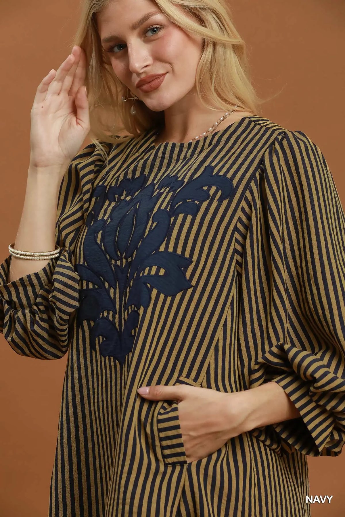 A woman wearing a navy blue and tan vertically striped tunic top with a large navy blue floral applique on the chest, posed against a solid warm brown background.