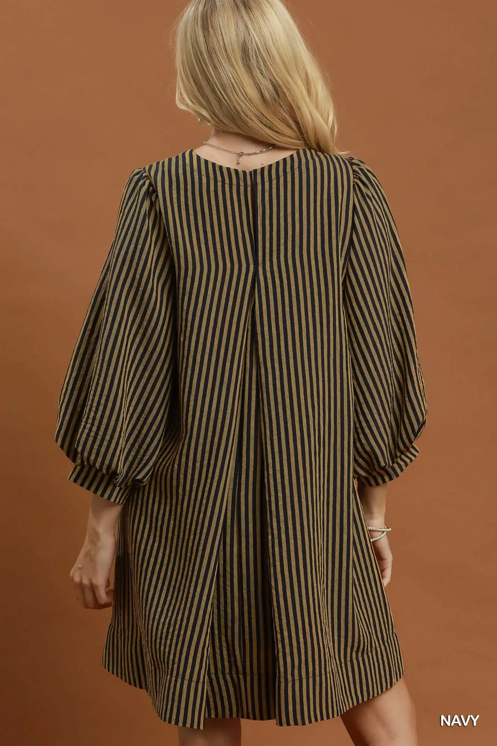 A woman shown from the back wearing a navy and olive green vertically striped dress with voluminous elbow-length sleeves, set against a solid warm brown studio background.