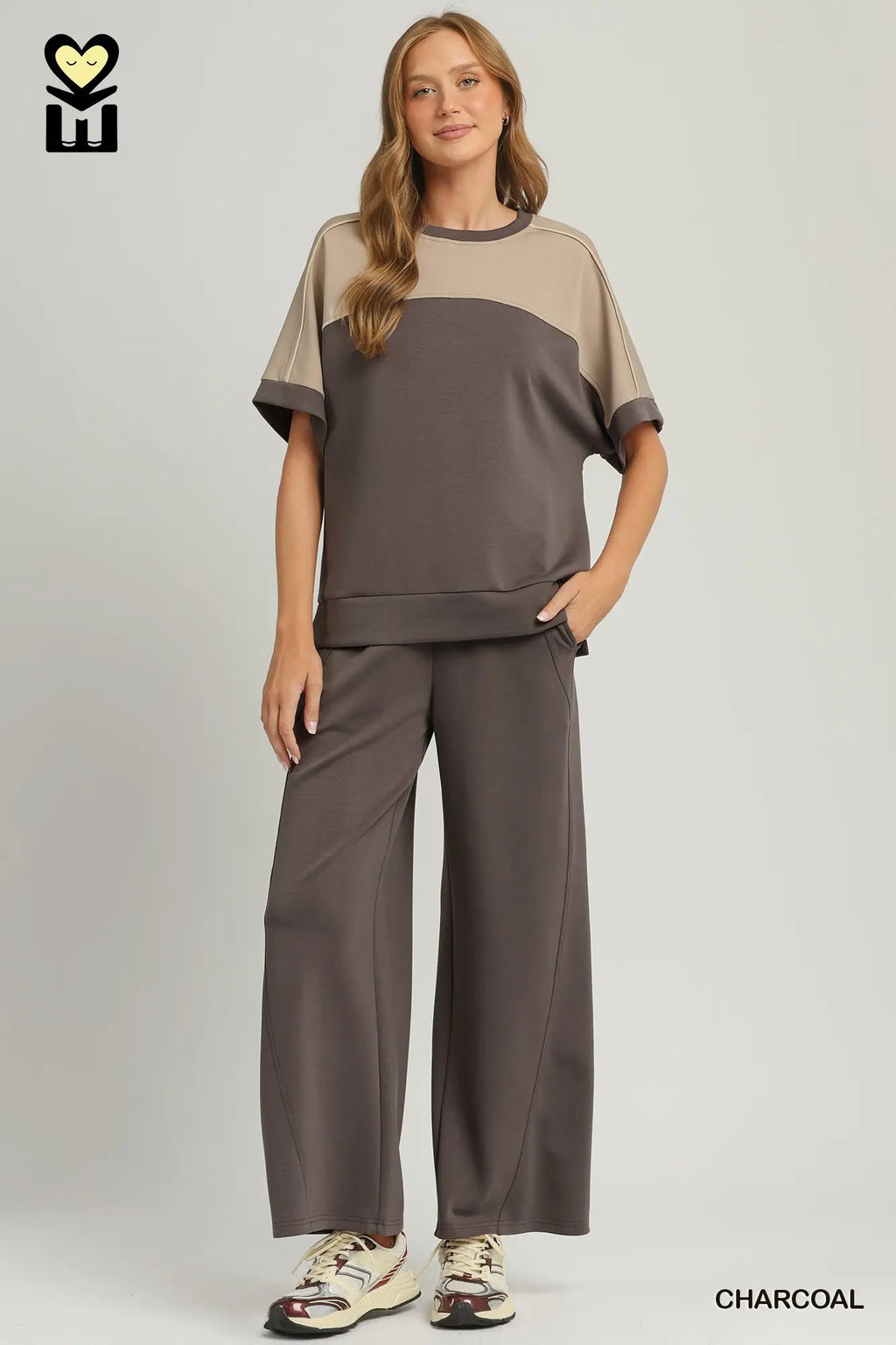 A woman modeling a two-piece lounge set in charcoal gray and beige. The set includes a short-sleeve top with a color-blocked design and matching wide-leg pants, shown against a light gray studio background.