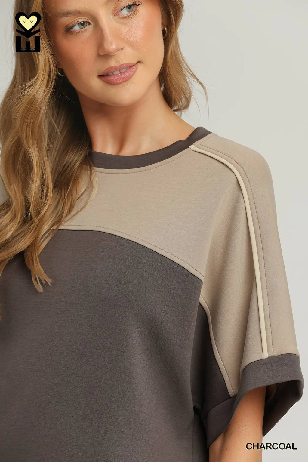 A woman wearing a color-block short-sleeve top in beige and charcoal gray, with a small black logo in the top-left corner and the word 'CHARCOAL' in the bottom-right corner.