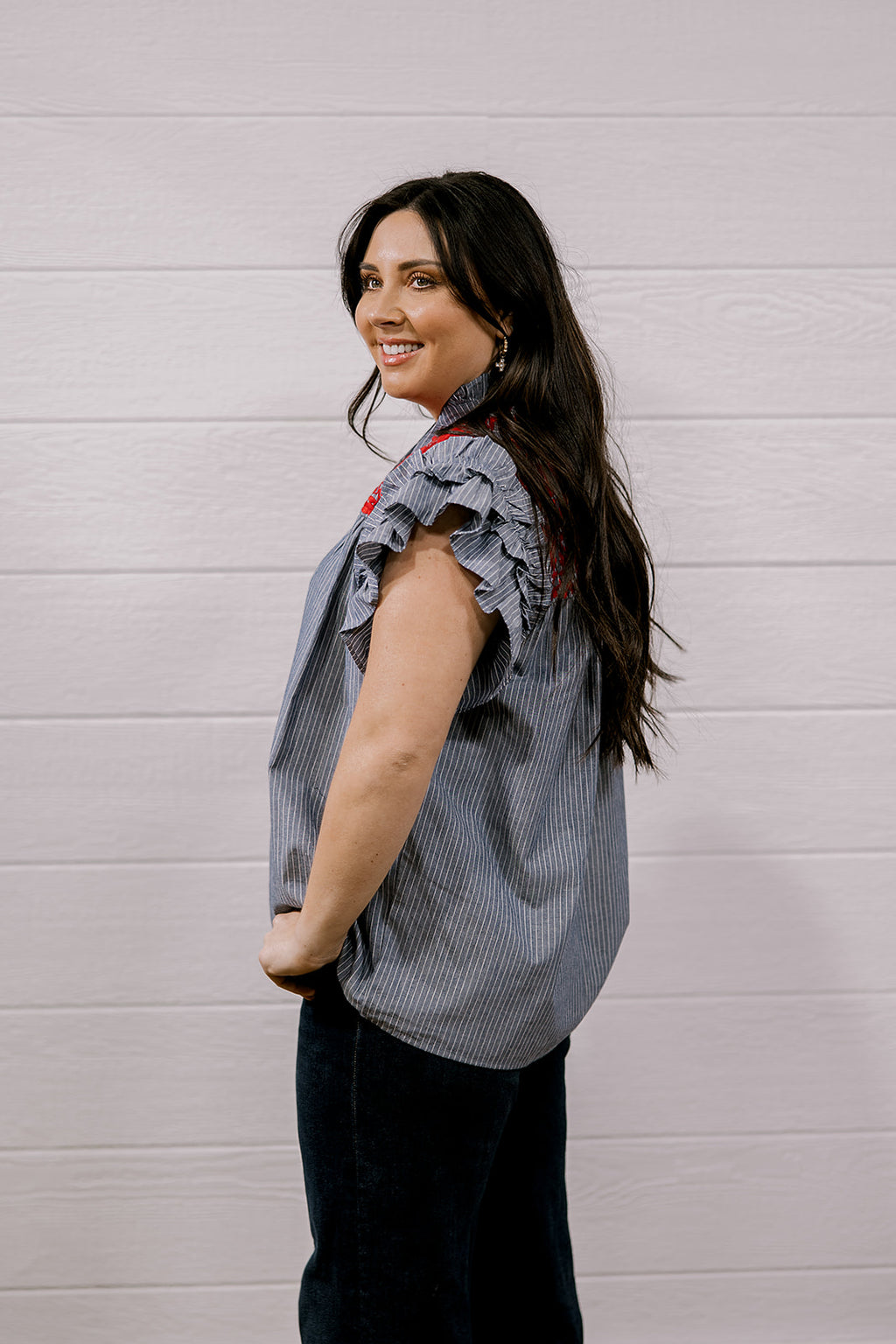 A woman with long dark hair is shown from the side, wearing a sleeveless ruffled blouse with a blue and white vertical stripe pattern and red accents, paired with dark pants, against a white paneled wall.