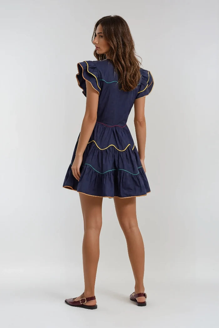A woman modeling a navy blue tiered mini dress from the back, featuring ruffled cap sleeves and colorful embroidered trim in yellow, green, and red, paired with dark brown flat sandals against a light gray studio background.
