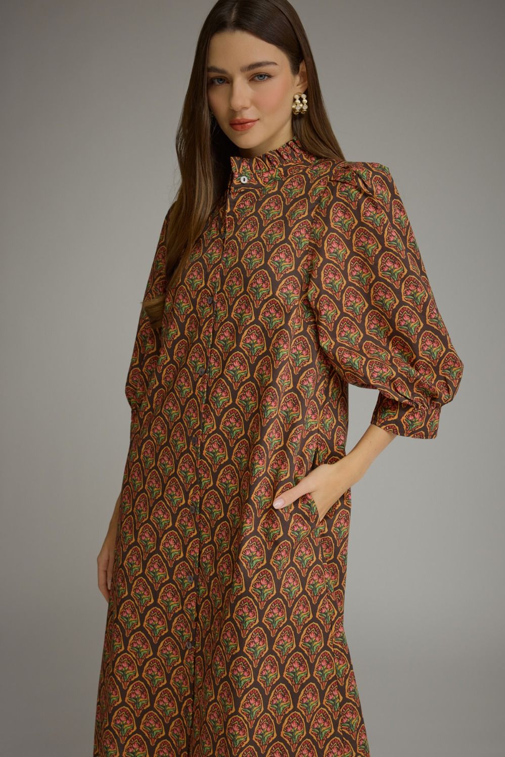 A woman modeling a long-sleeved midi dress with a high mock neck and voluminous elbow-length puff sleeves. The dress features an all-over repeating paisley pattern in brown, rust red, and olive green.