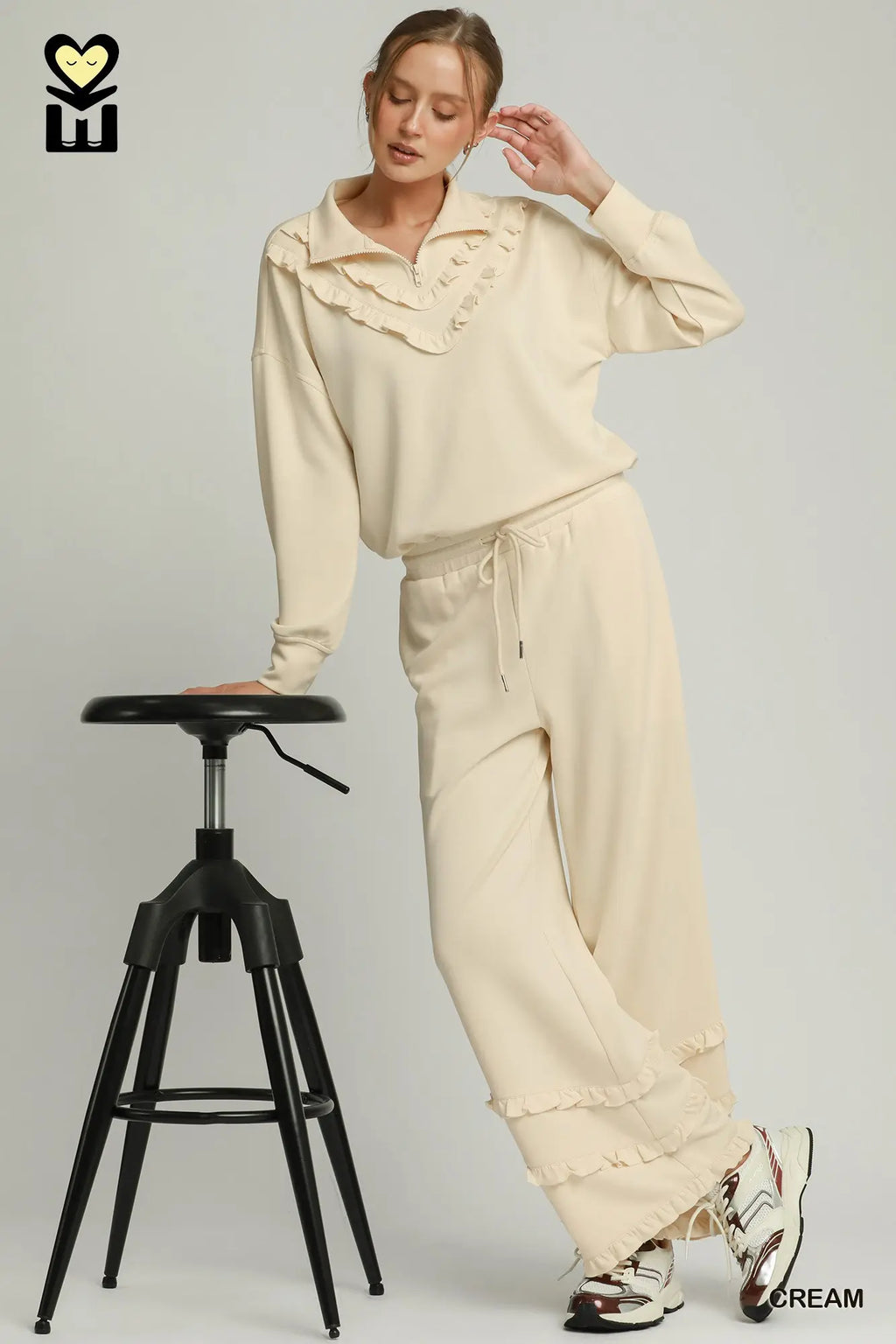 Woman in beige outfit standing next to a black stool on a plain background