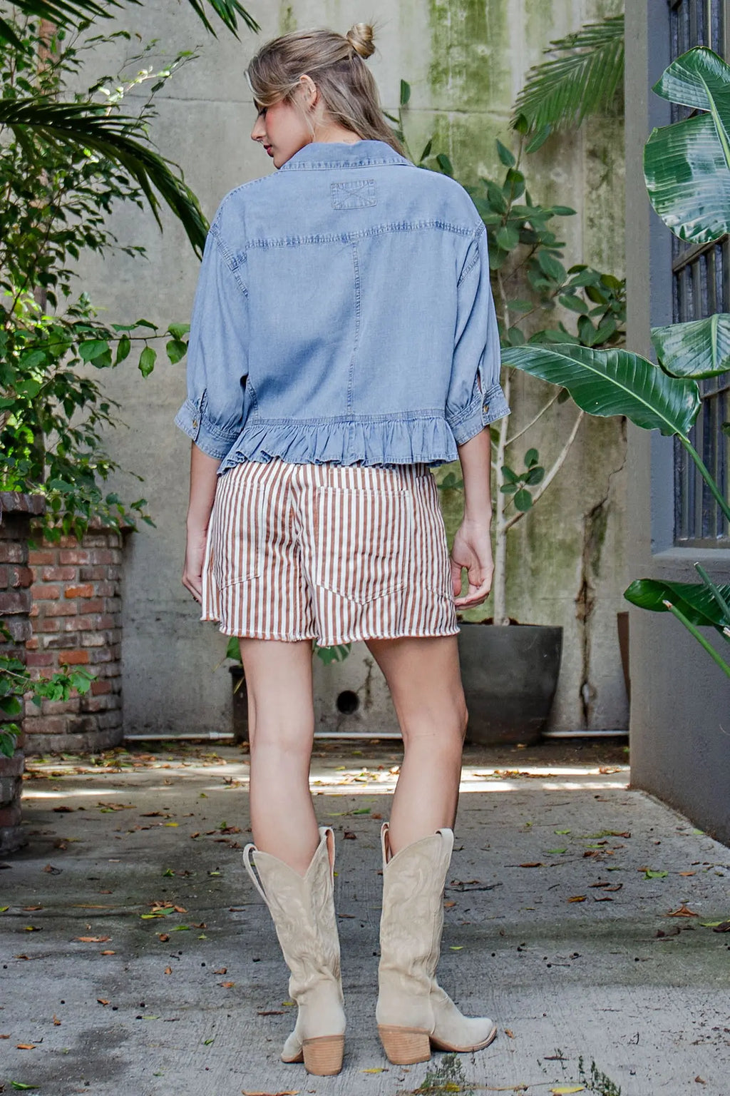 Woman wearing a denim jacket, striped shorts, and beige cowboy boots standing outdoors.
