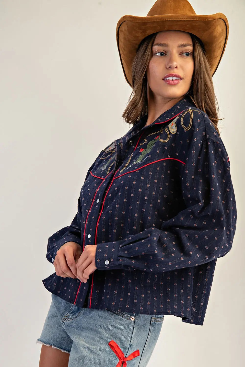 Woman wearing a navy patterned shirt, denim shorts, and a brown cowboy hat on a plain background