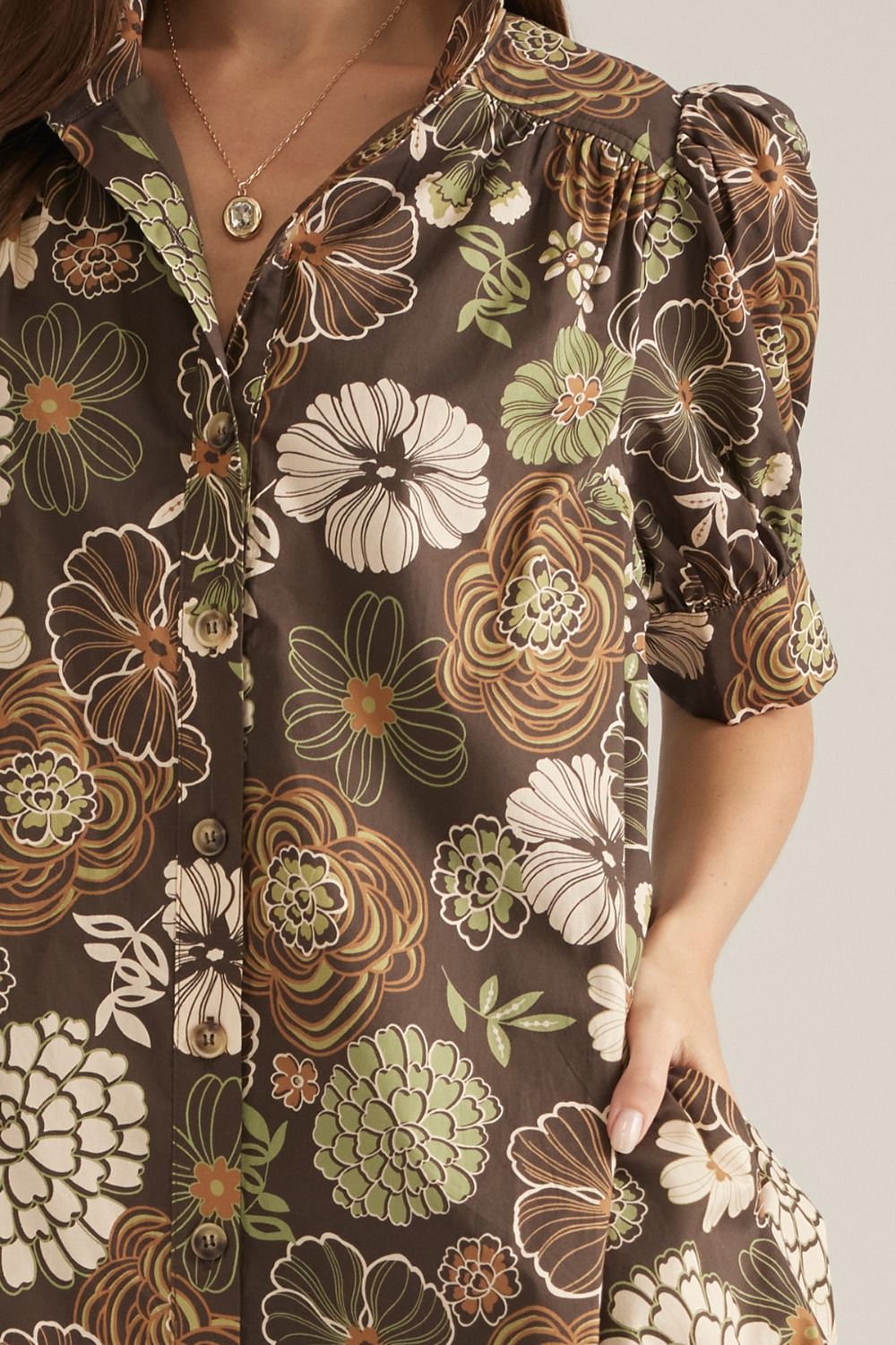 A close-up of a woman wearing a brown short-sleeve button-up shirt with a retro floral print in beige, white, and olive green. The shirt features puffed short sleeves and a button-down front.
