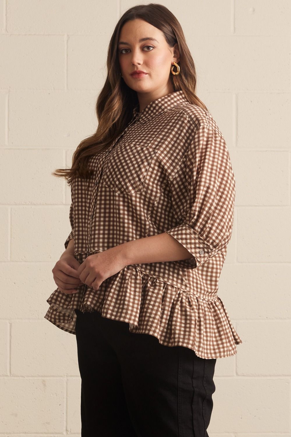 Woman wearing a brown and white checkered blouse with ruffled hem against a beige brick wall.