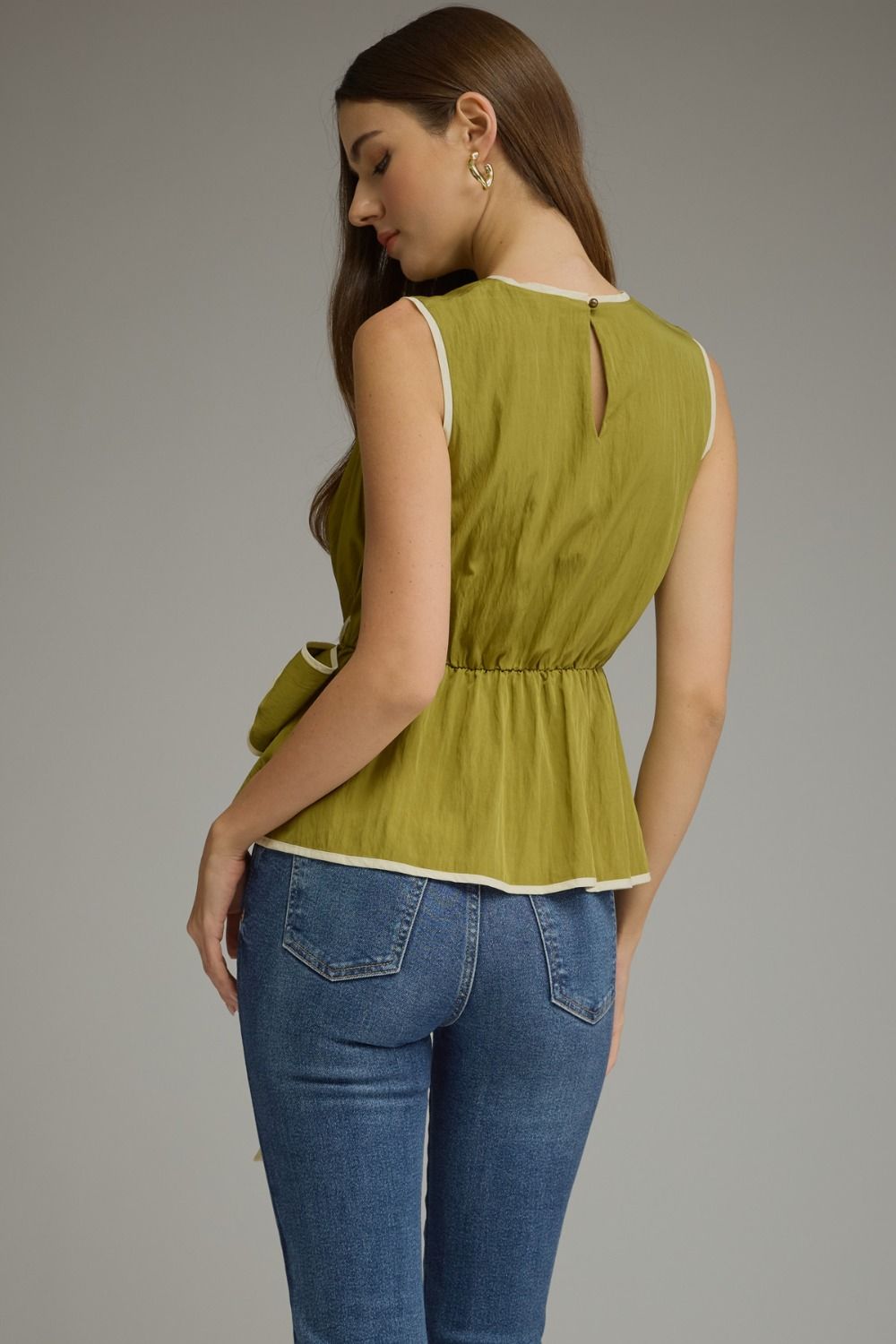 A woman shown from the back wearing a sleeveless olive green peplum top with cream trim and blue jeans, photographed against a plain gray studio background.