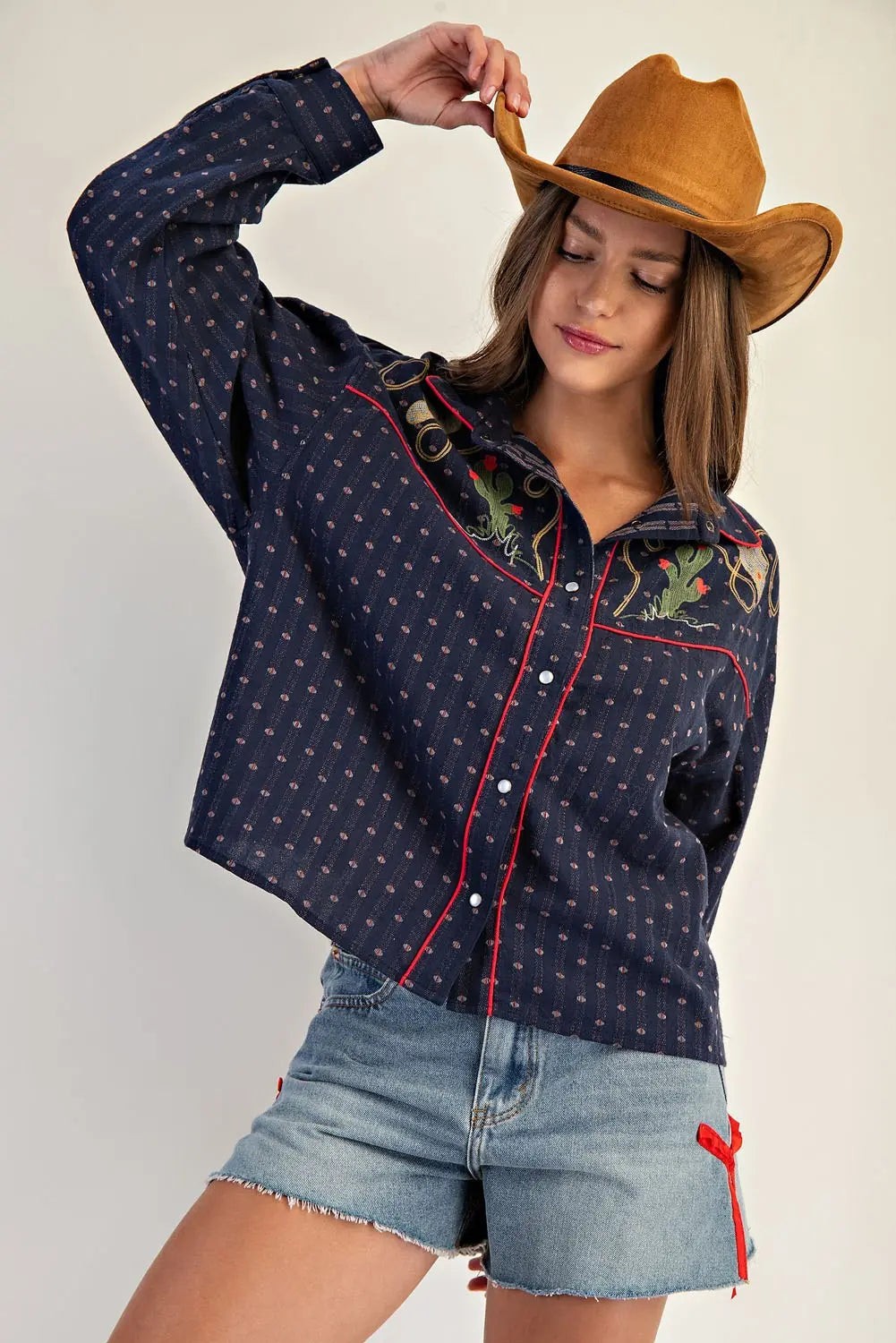 Woman wearing a patterned shirt and denim shorts, holding a cowboy hat against a plain background