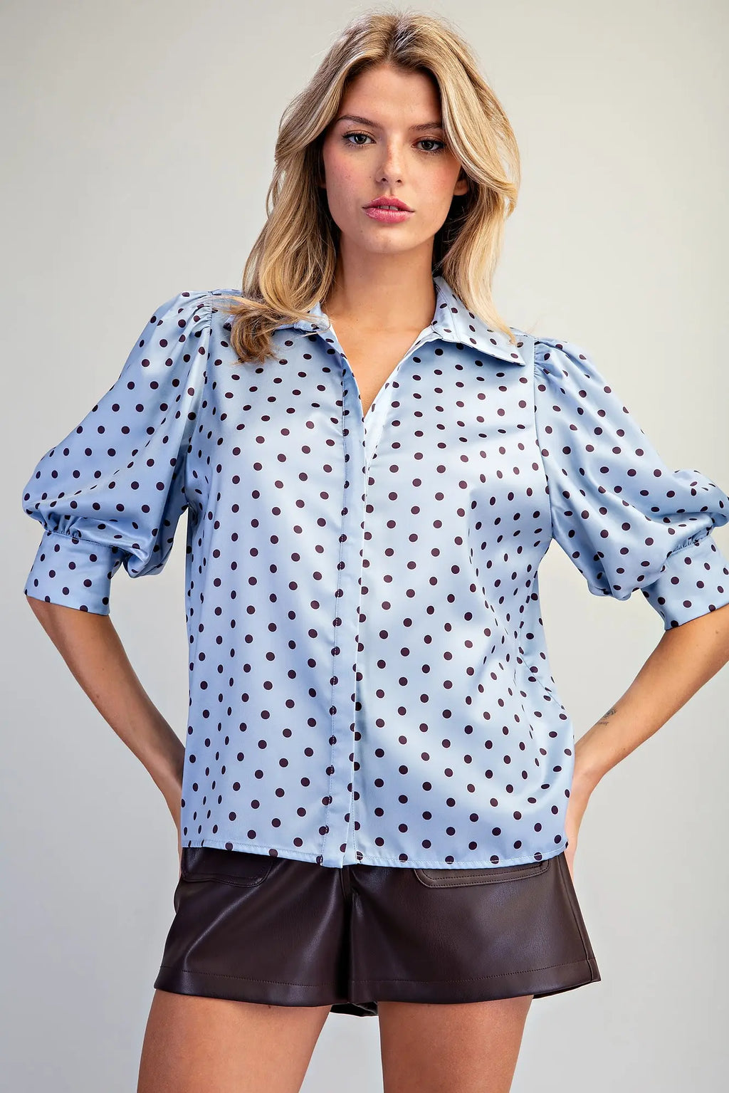 A woman modeling a light blue button-up blouse with black polka dots and puffed sleeves, paired with dark brown shorts, against a plain light gray studio background.