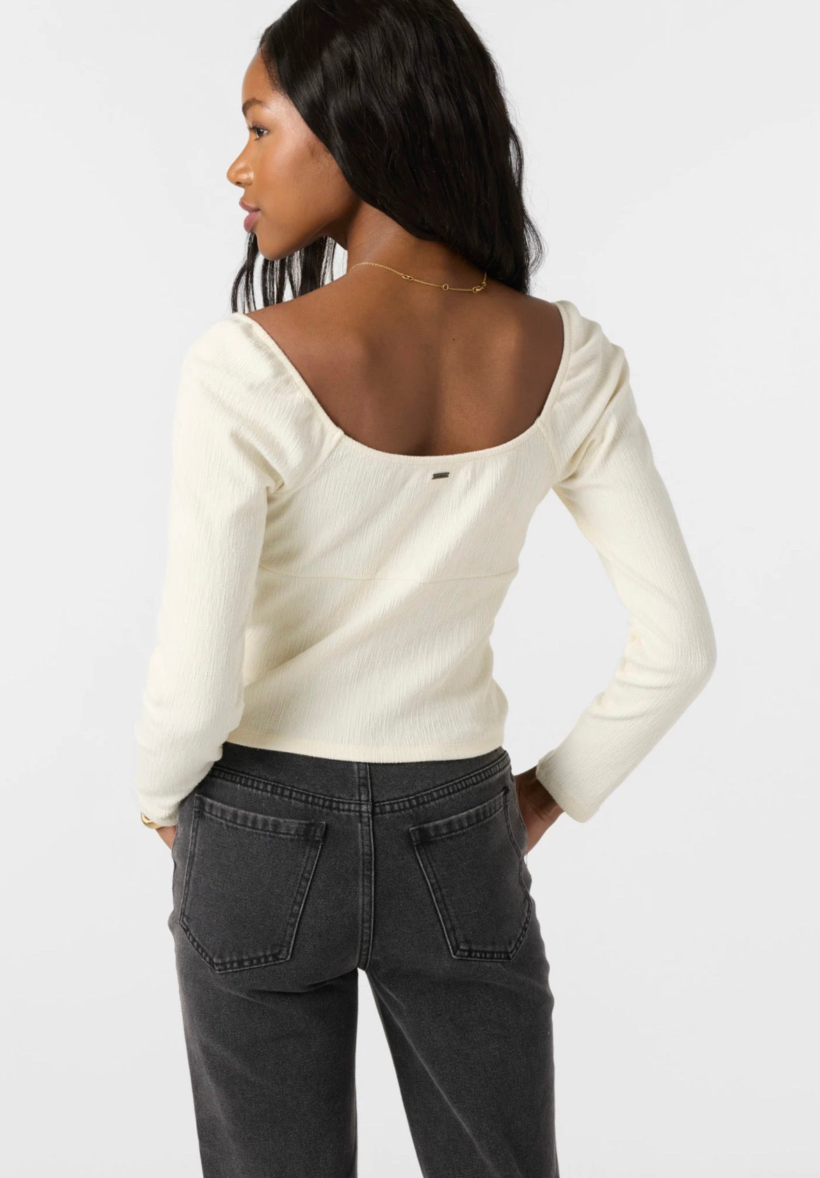 A woman shown from the back wearing a cream ribbed long-sleeve top with a wide scoop neckline and dark gray jeans, against a plain light gray studio background.