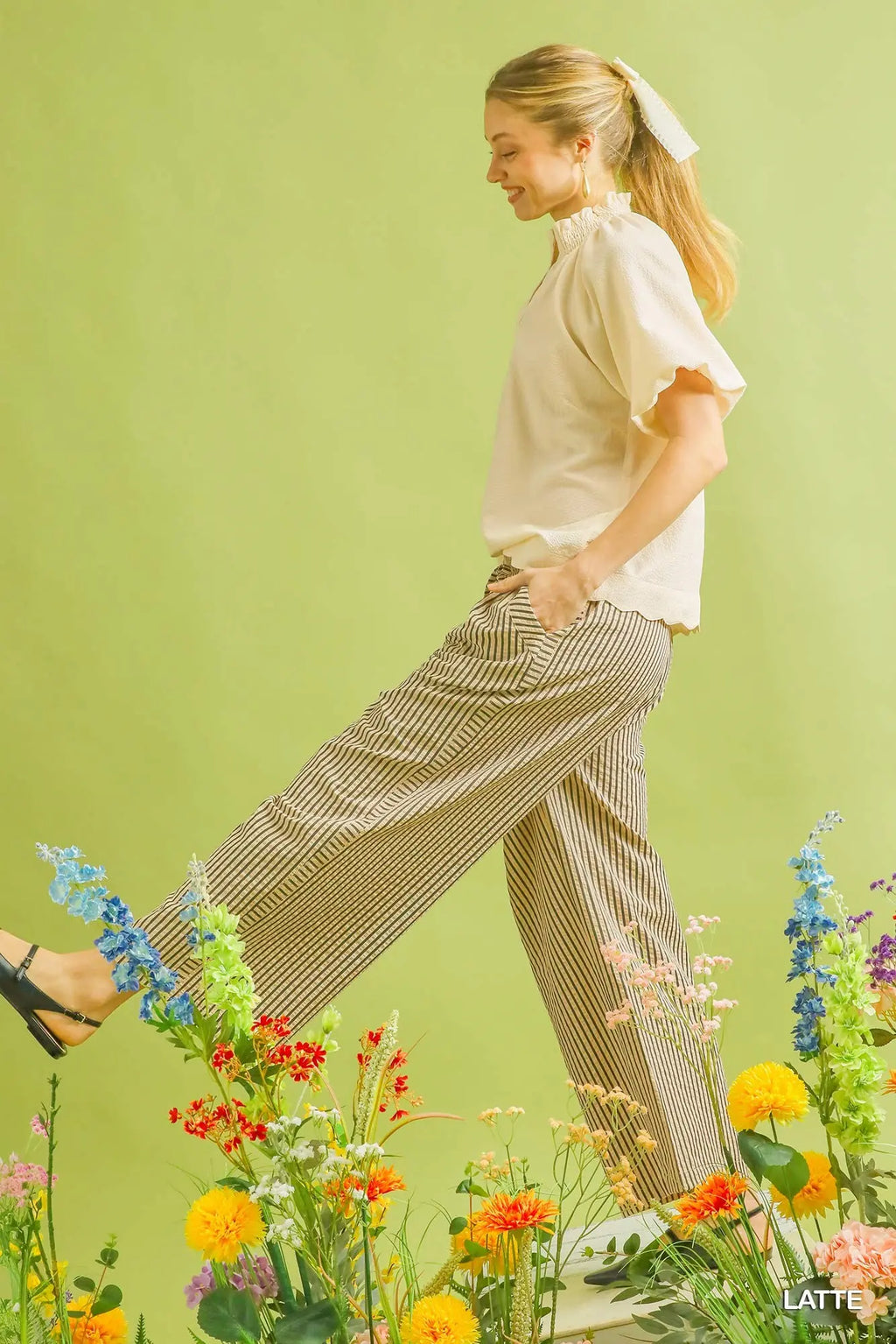 Woman in a white blouse and patterned pants standing among flowers against a green background