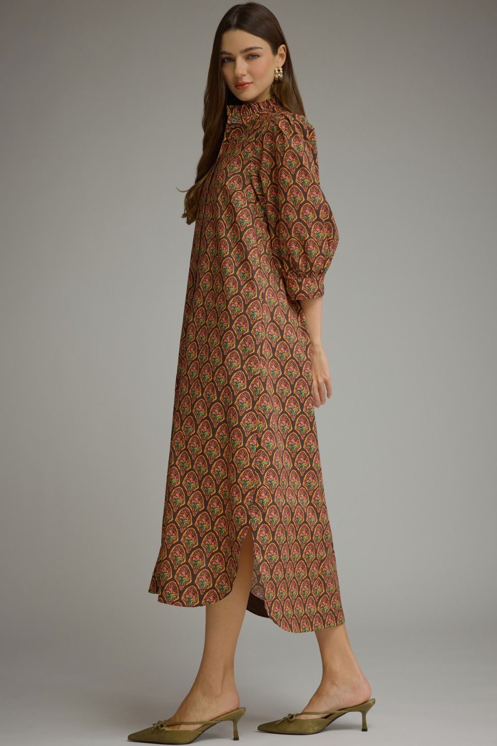 A woman modeling a long-sleeved midi dress with a brown, red, and green scalloped pattern, paired with olive green pointed-toe mule heels, set against a plain gray studio background.