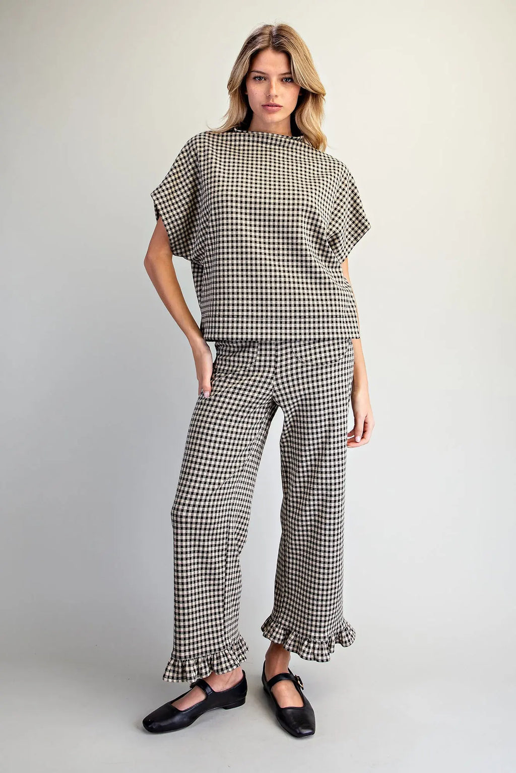 A woman modeling a matching two-piece outfit featuring a black and white gingham patterned short-sleeve top and wide-leg pants with ruffled hems, paired with black Mary Jane shoes, against a light gray studio background.