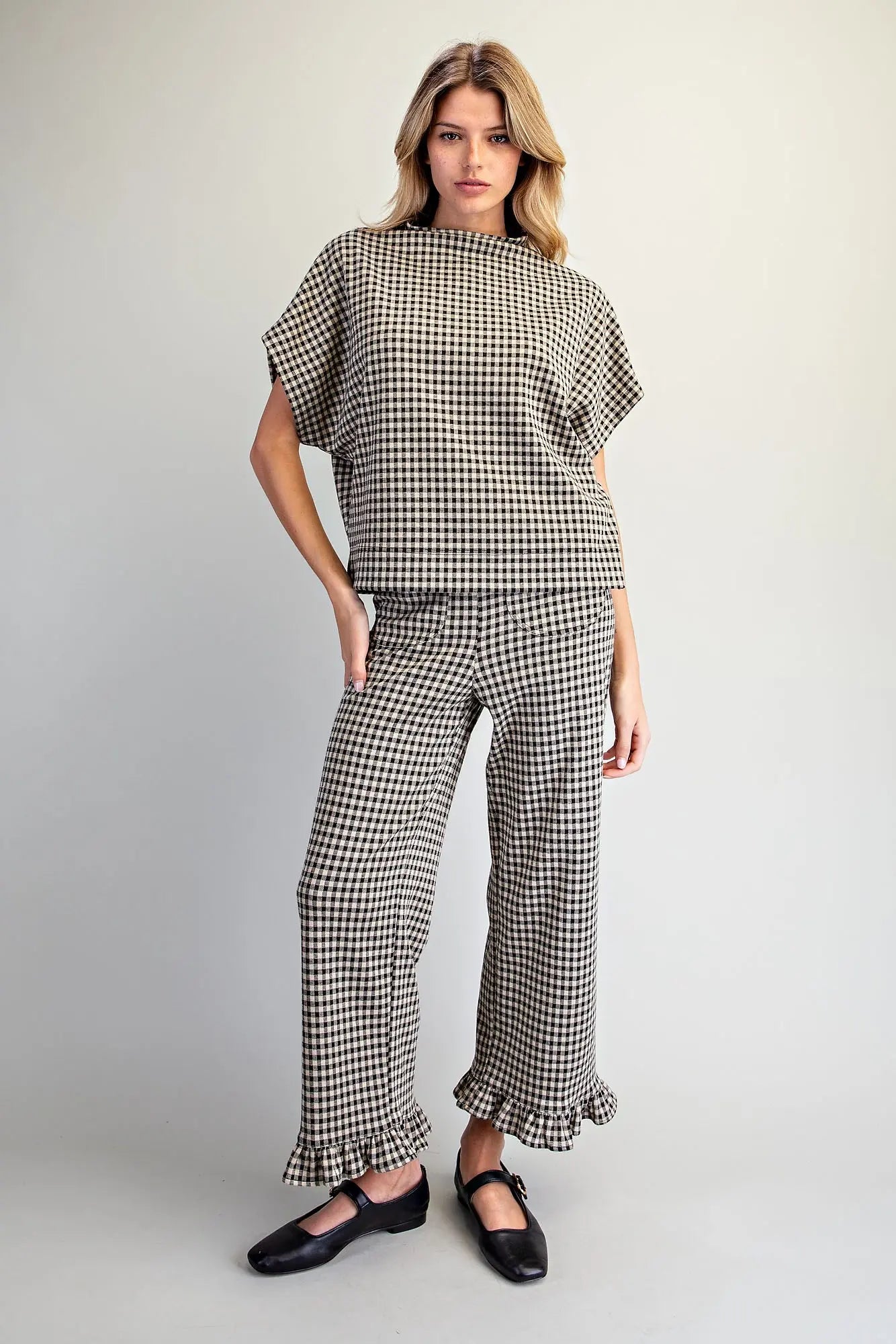 A woman modeling a matching two-piece outfit featuring a black and white gingham patterned short-sleeve top and wide-leg pants with ruffled hems, paired with black Mary Jane shoes, against a light gray studio background.