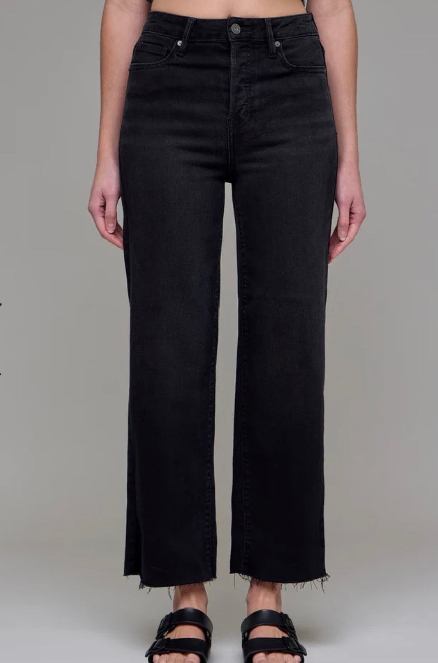 A model wearing a pair of black wide-leg cropped jeans with a raw, frayed hem, paired with black sandals, shown against a neutral gray studio background.