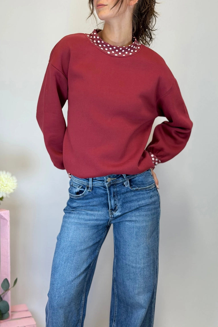 A woman wearing a burgundy crewneck sweatshirt with a white polka-dot collar and matching polka-dot cuffs, paired with blue jeans.