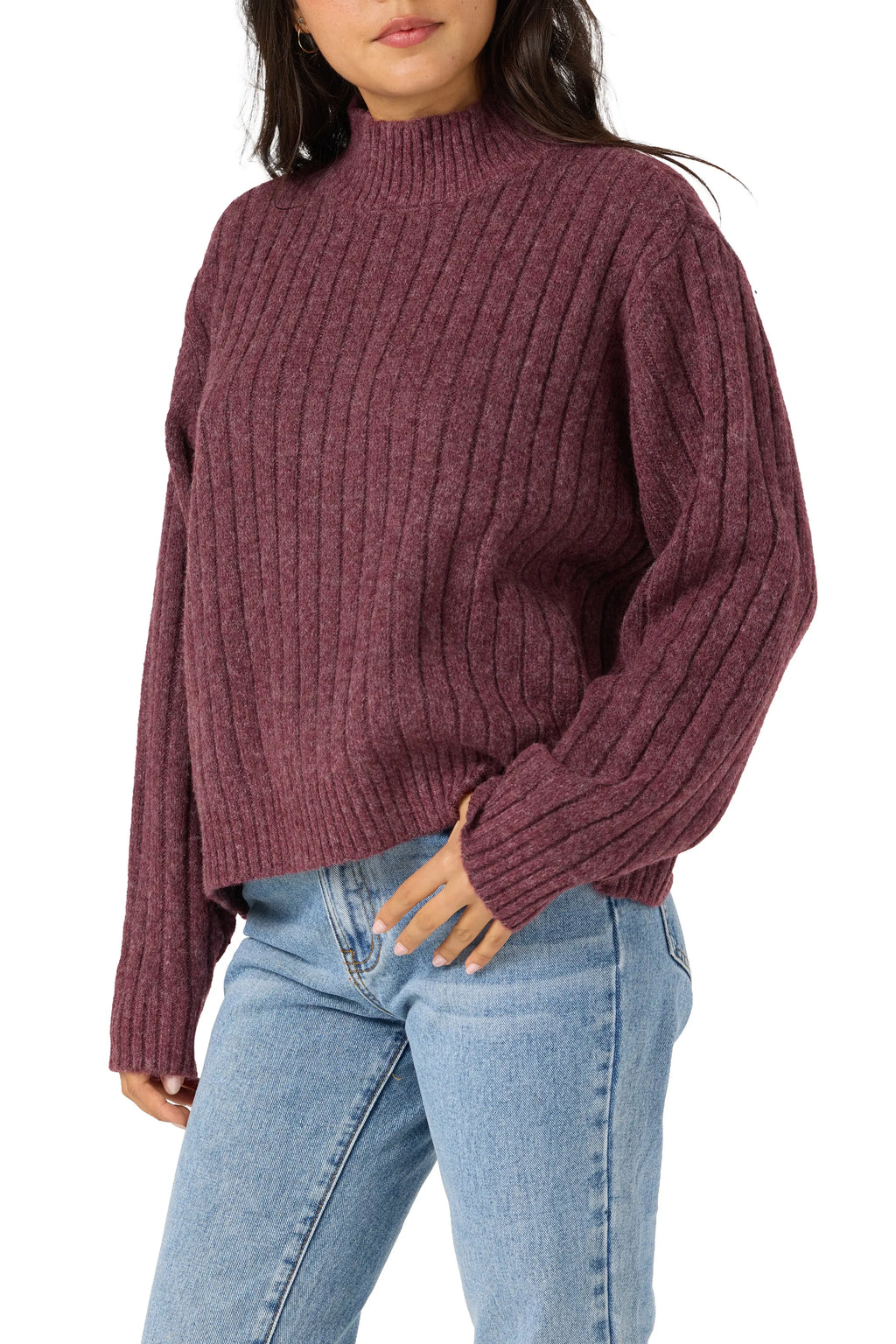 A woman wearing a ribbed knit mock neck sweater in a dusty plum purple color, paired with light blue jeans, against a plain white background.