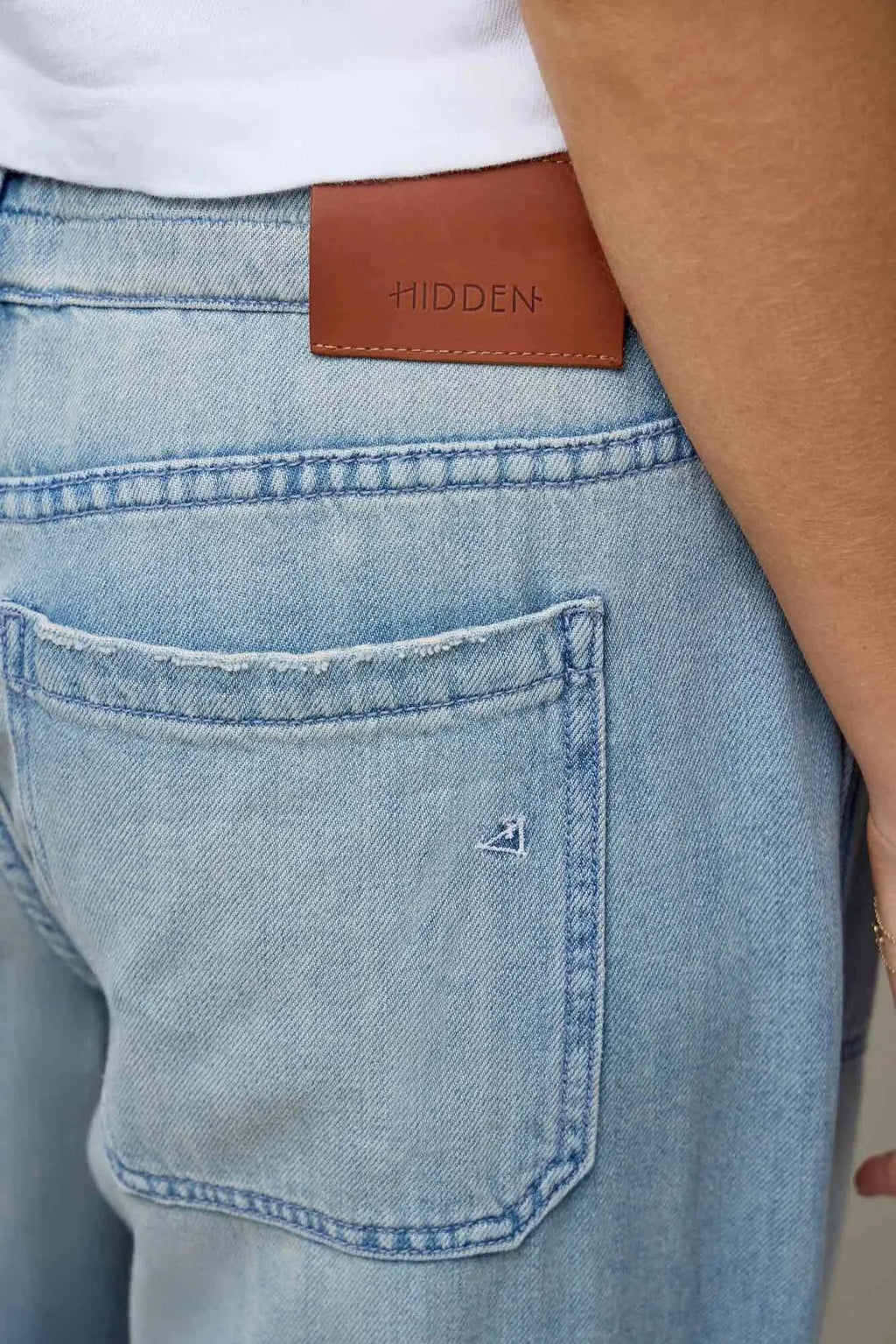 Close-up of a person wearing light-wash blue denim jeans with a brown leather patch at the back waistband embossed with the brand name 'HIDDEN'.