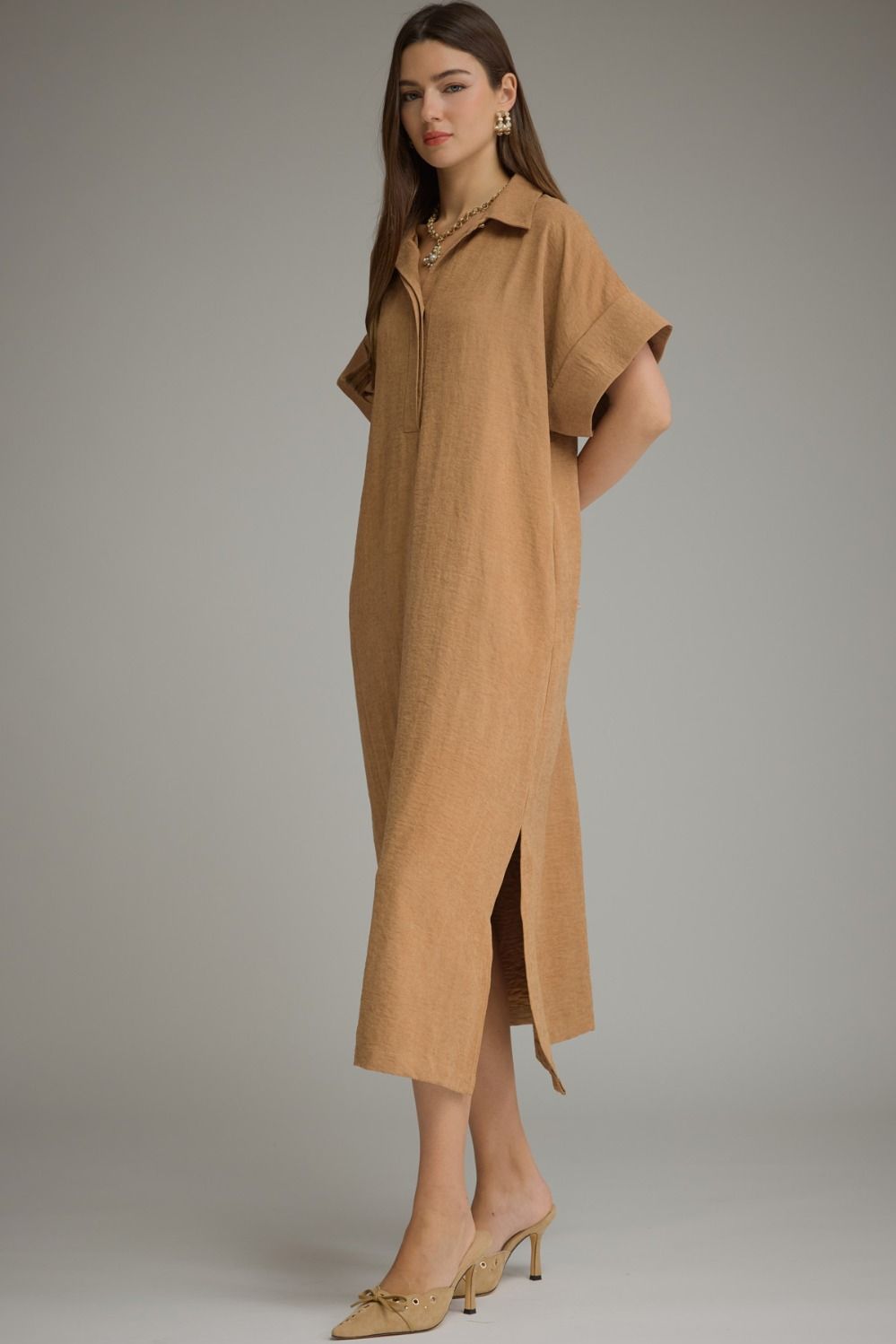 A woman modeling a tan linen shirt dress with short flutter sleeves, a button-down collar, and a side slit, paired with beige pointed-toe heels against a neutral gray studio background.