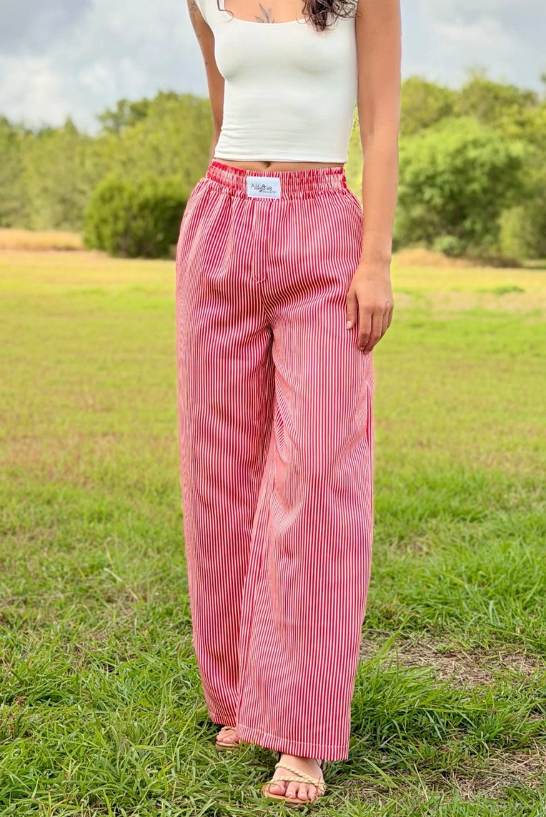 A person standing outdoors on grass wearing loose, wide-leg pink striped pants with an elastic waistband and a white ribbed tank top.
