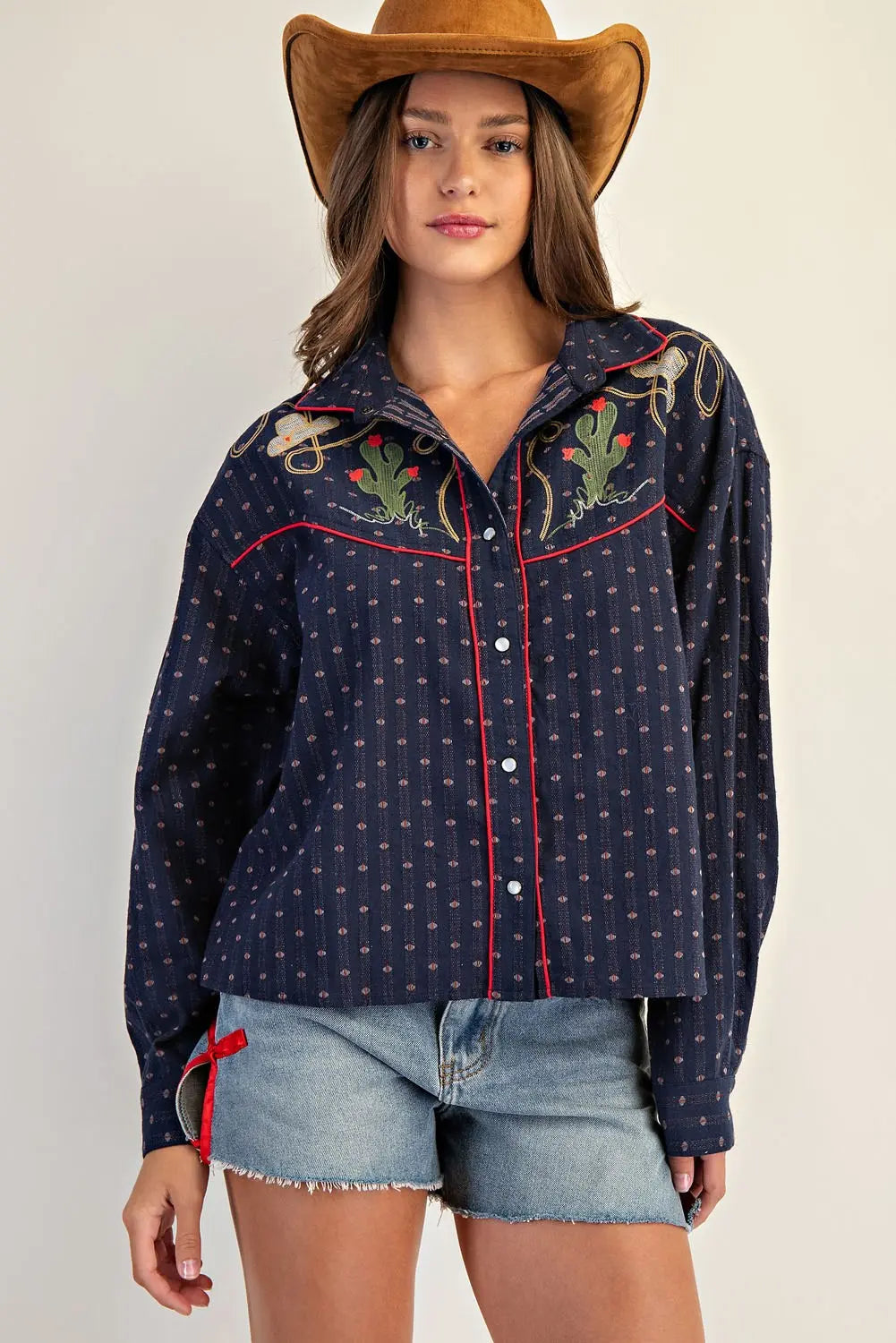 Woman wearing a navy blue embroidered shirt with red accents and denim shorts on a plain background
