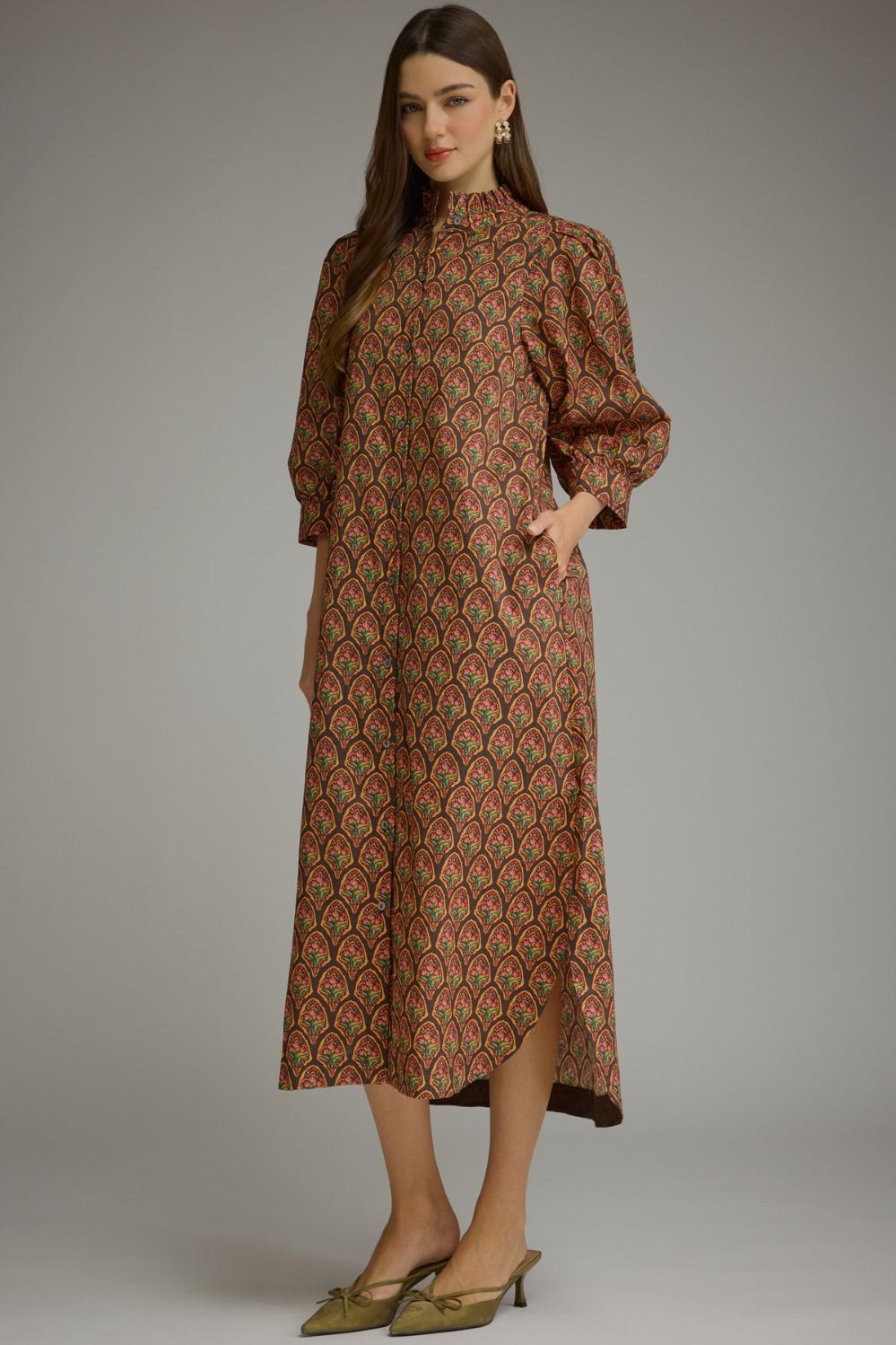 A woman modeling a long-sleeved midi dress with a brown base and a repeating red, green, and gold scalloped pattern, paired with olive green pointed-toe mule heels against a neutral gray studio background.