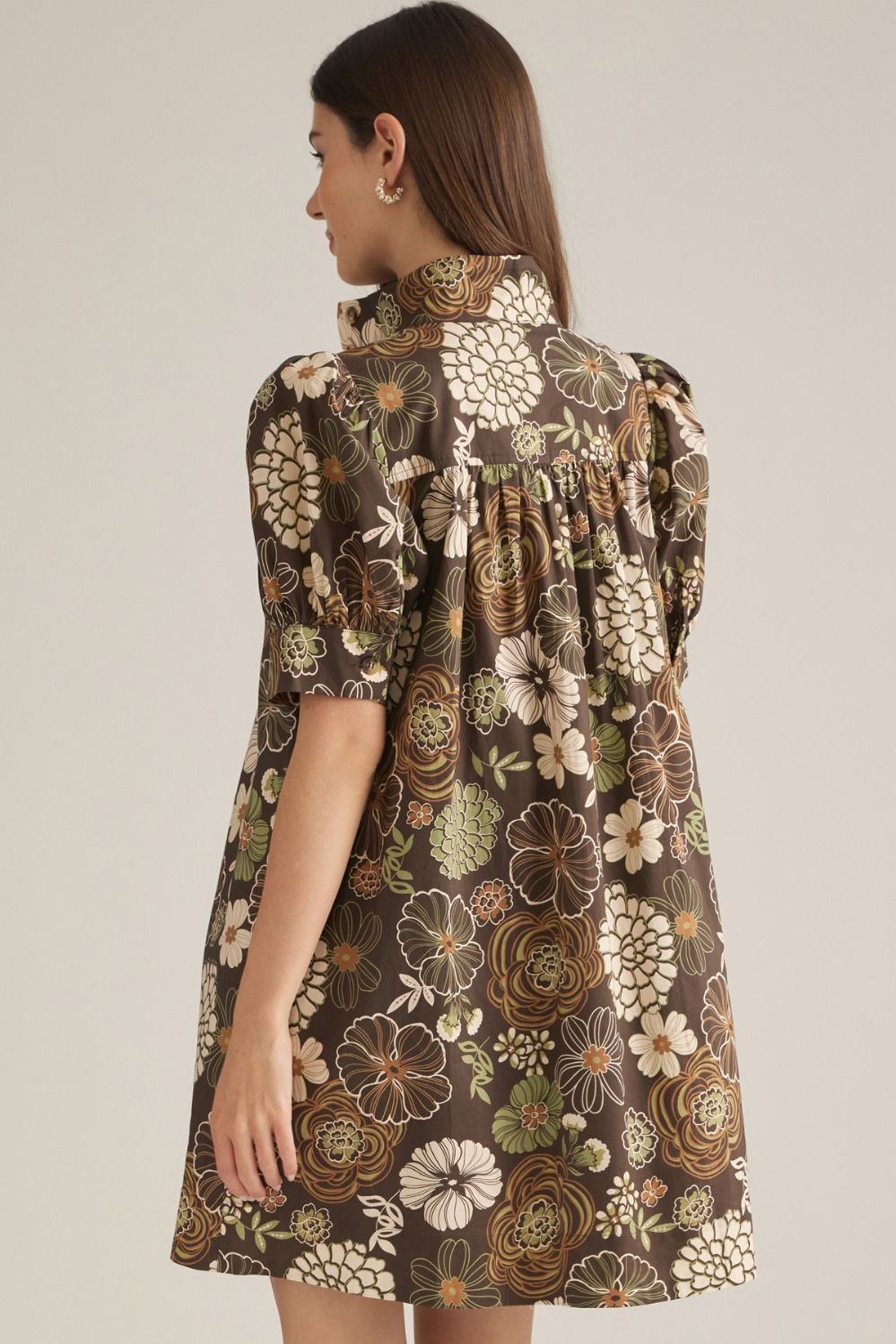 A woman shown from the back wearing a brown short-sleeve dress with a dense floral pattern in beige, white, and muted green, against a plain light gray studio background.