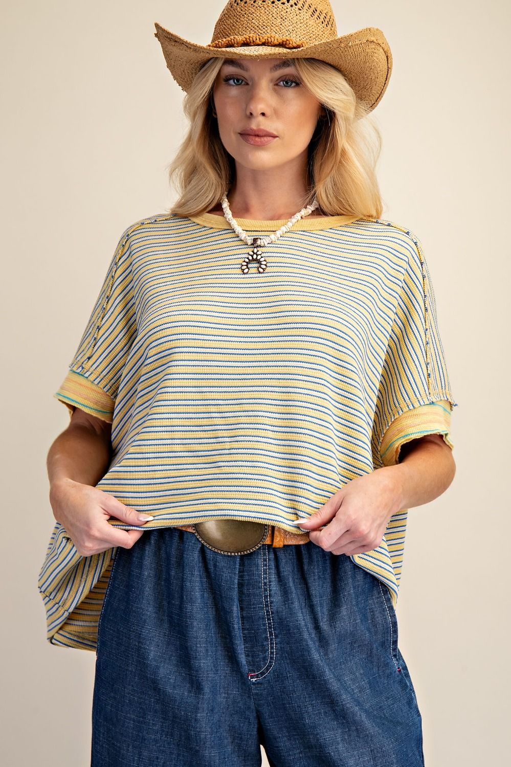 Woman wearing a striped shirt, cowboy hat, and blue jeans against a beige background