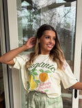 Woman wearing a 'organic' t-shirt with fruit graphics and green shorts, standing in front of a glass door.
